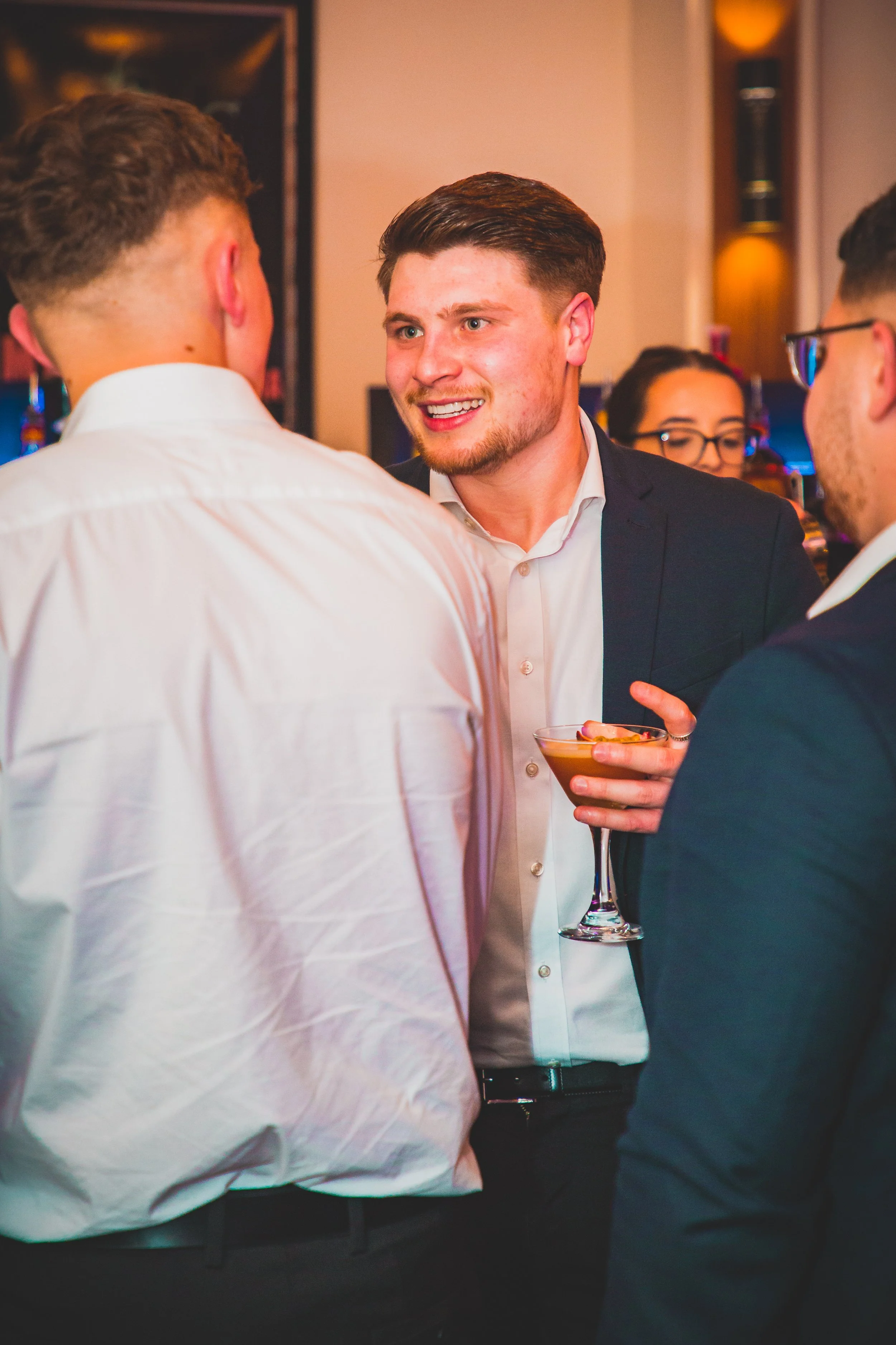 Four young men dressed in formal attire are engaged in conversation at a social gathering, with one holding a cocktail glass.