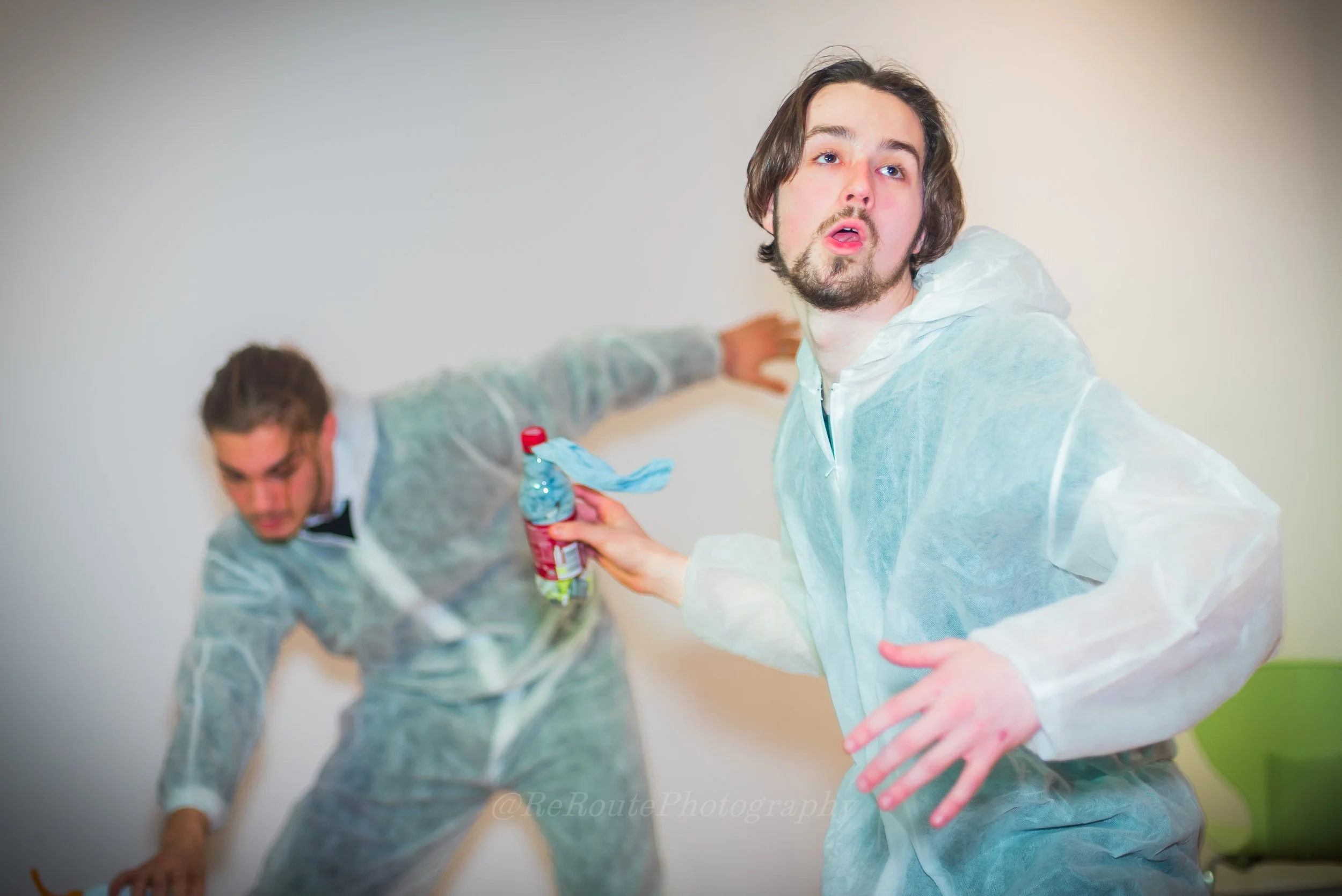 Two young men in gray hoodies engaged in a playful pillow fight, one holding a water bottle, inside a room with plain walls.