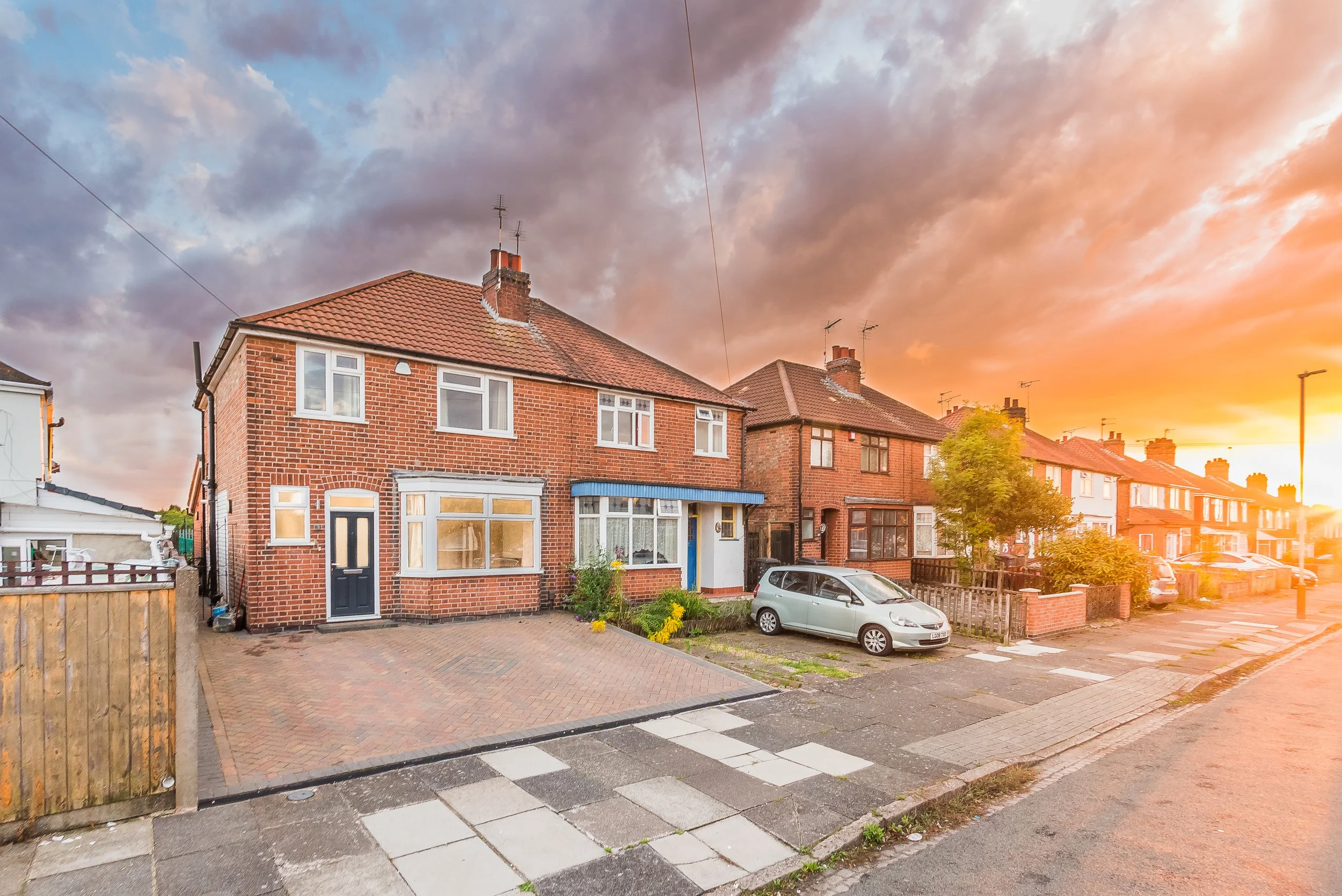 A row of brick houses on a residential street during sunset with a parked car and a tree in front.