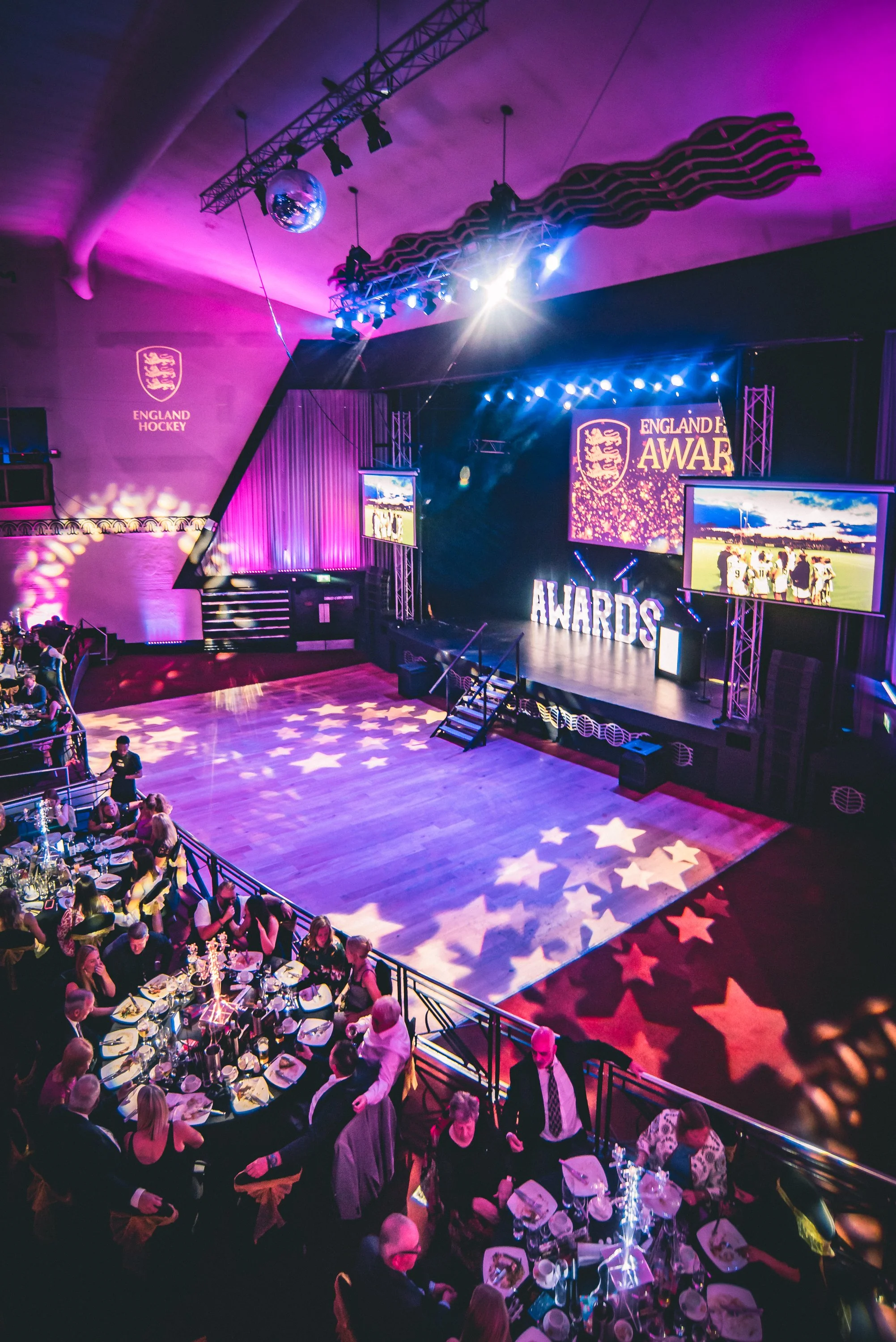 An awards ceremony event with people dining at round tables, stage with large screens displaying 'England Hockey Awards', starry lighting on the dance floor, purple and pink lighting, and a disco ball hanging from the ceiling.
