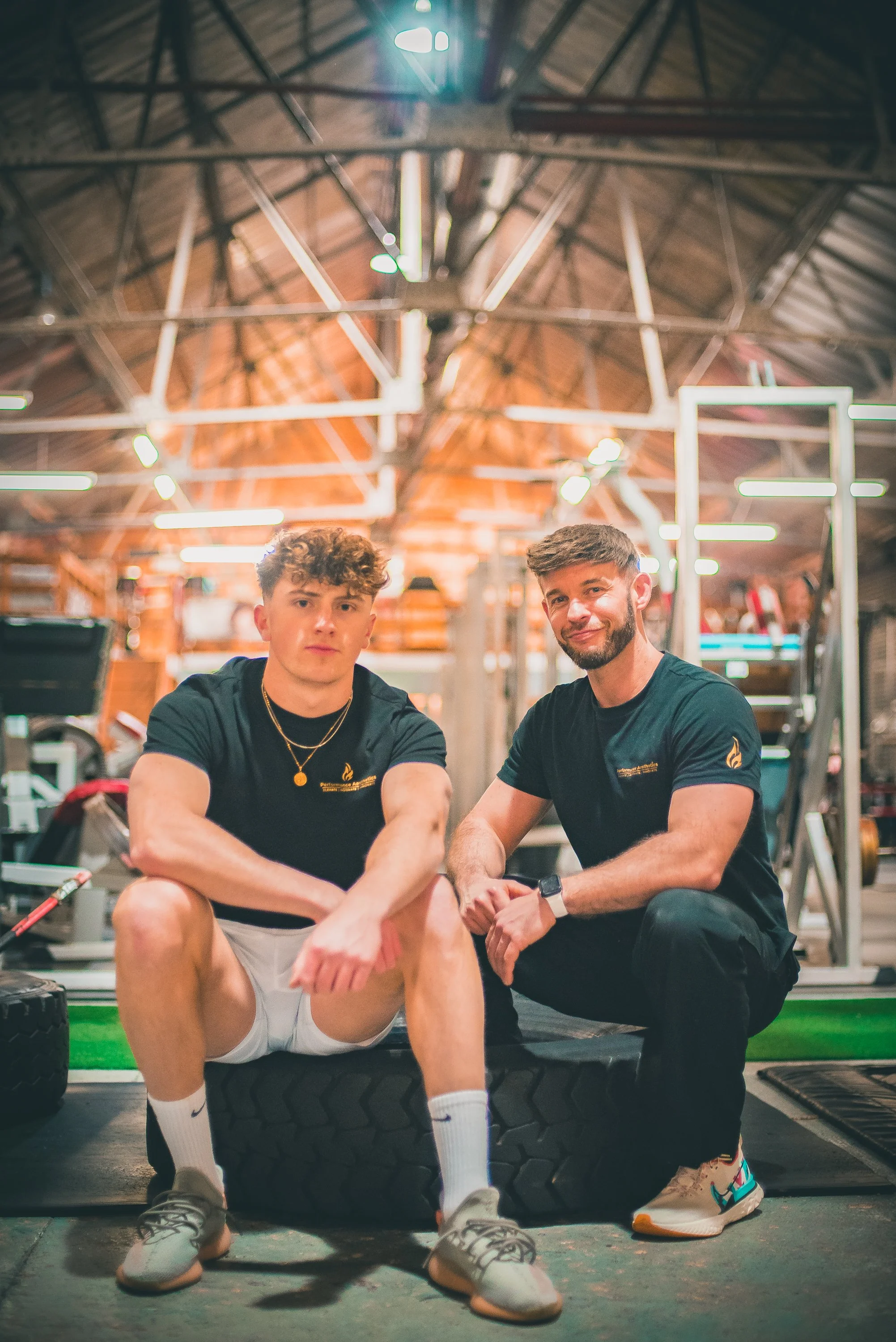 Two young men in athletic clothing sitting in a gym with a wooden ceiling and bright lighting. One man has curly hair and is wearing a gold chain, while the other has short hair and a beard. They are sitting on a large tire with gym equipment around 