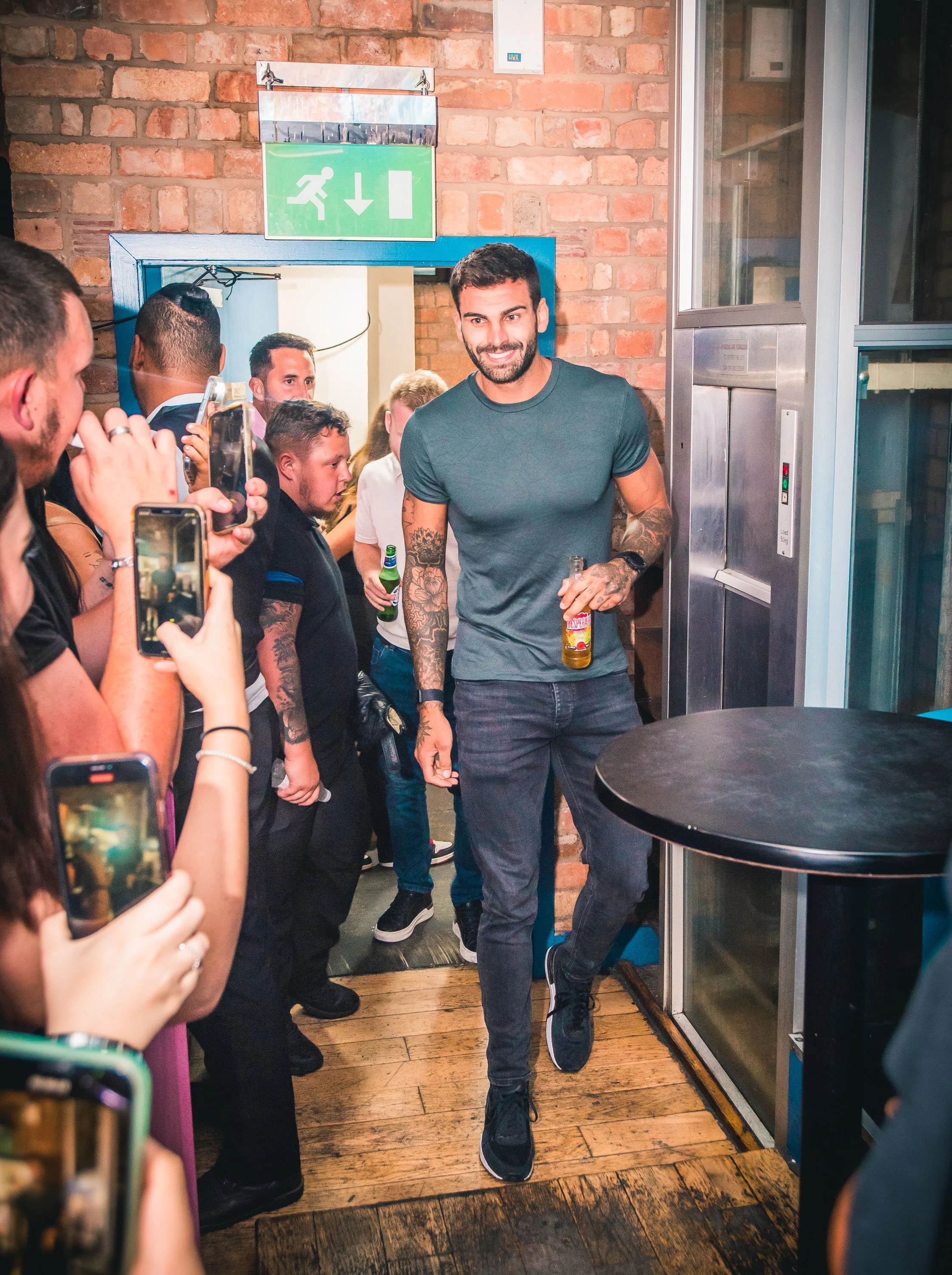 A man with tattoos on his arms smiling while holding a drink, walking through a crowd in what appears to be a bar or club. The crowd is taking photos and videos of him. The background has a brick wall and an emergency exit sign.