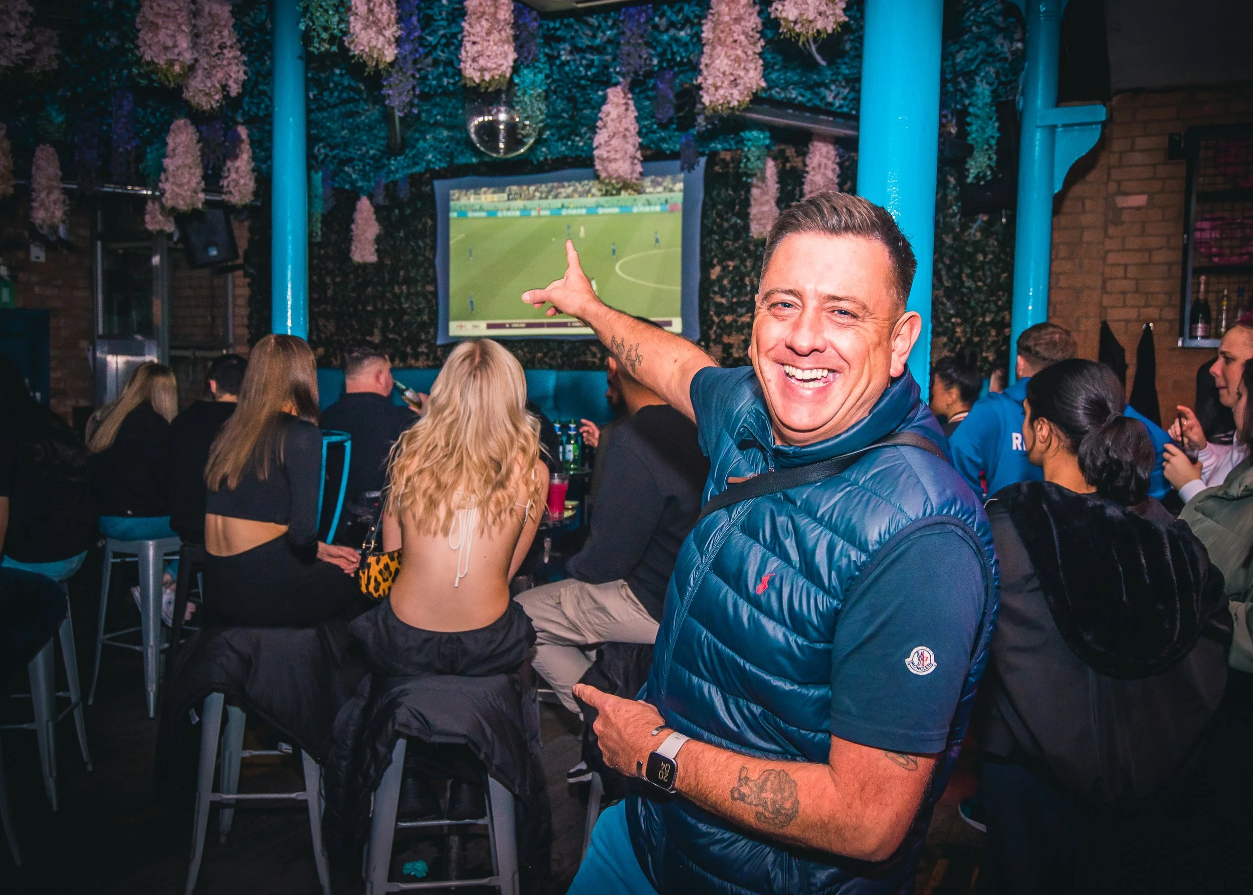 A man smiling and pointing towards a television screen showing a soccer match, in a lively bar filled with other people watching the game.