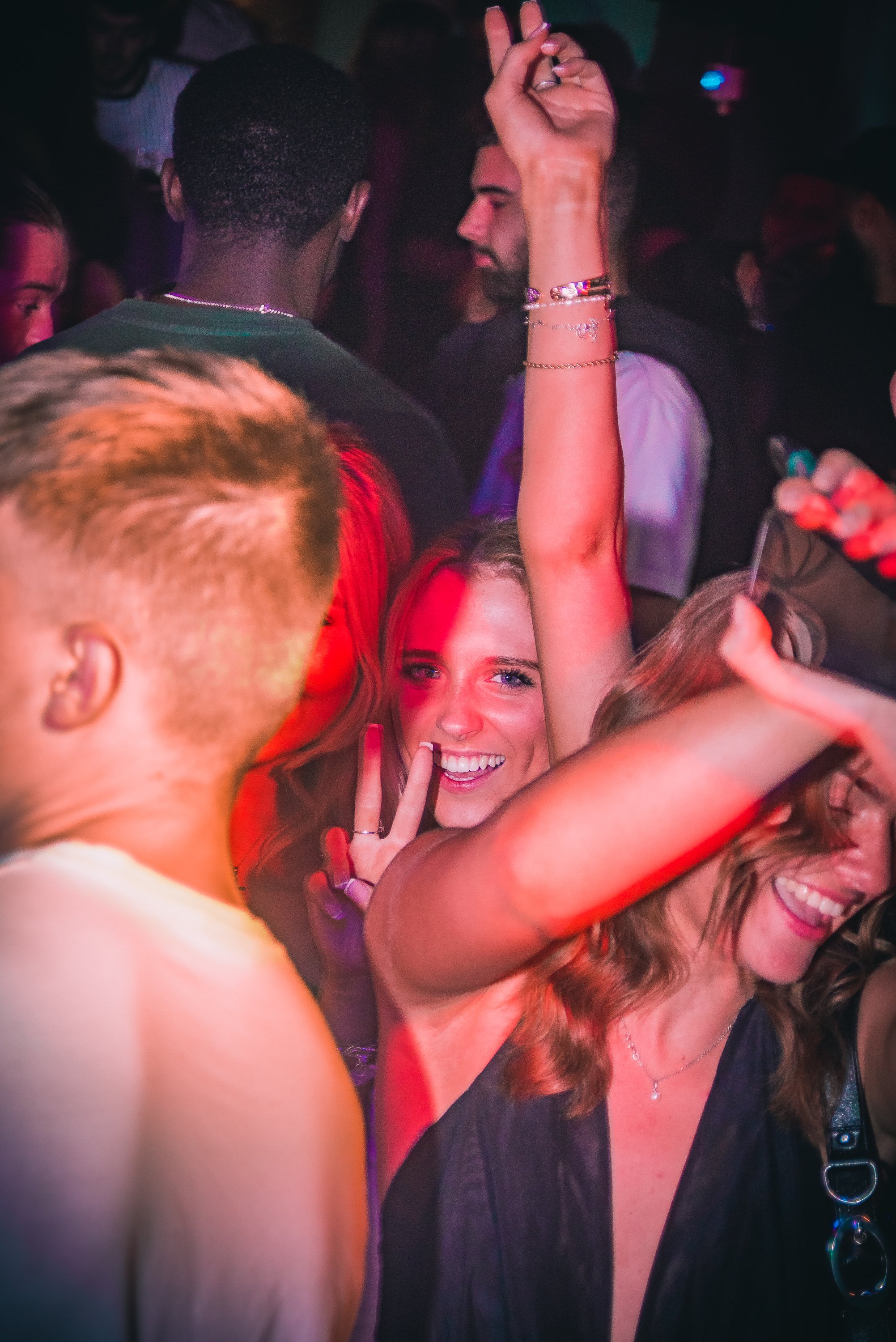 People dancing and enjoying themselves at a lively party or nightclub, with one woman making a peace sign and smiling brightly.