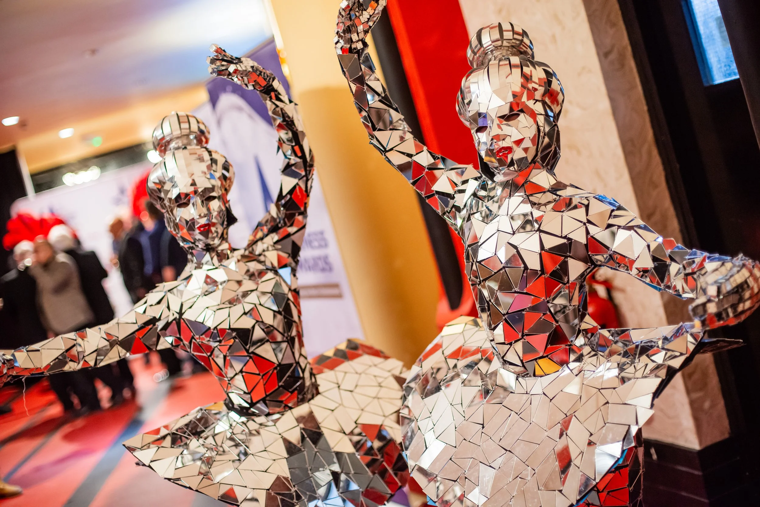 A reflective sculpture of a reindeer made of small, triangular mirror pieces, displayed indoors at an event with several people in the background.