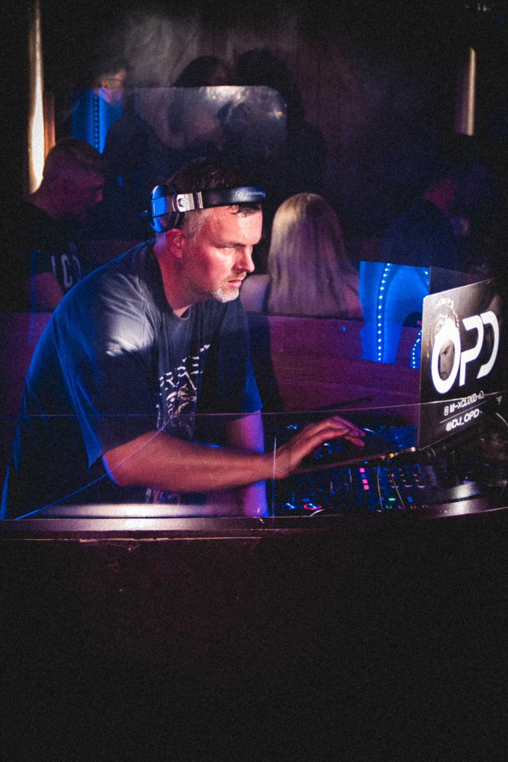 A man wearing a black t-shirt and headphones is sitting at a DJ booth in a dimly lit club or event space, with purple and blue lighting. There are people in the background and a sign with the word 'OP' partially visible.