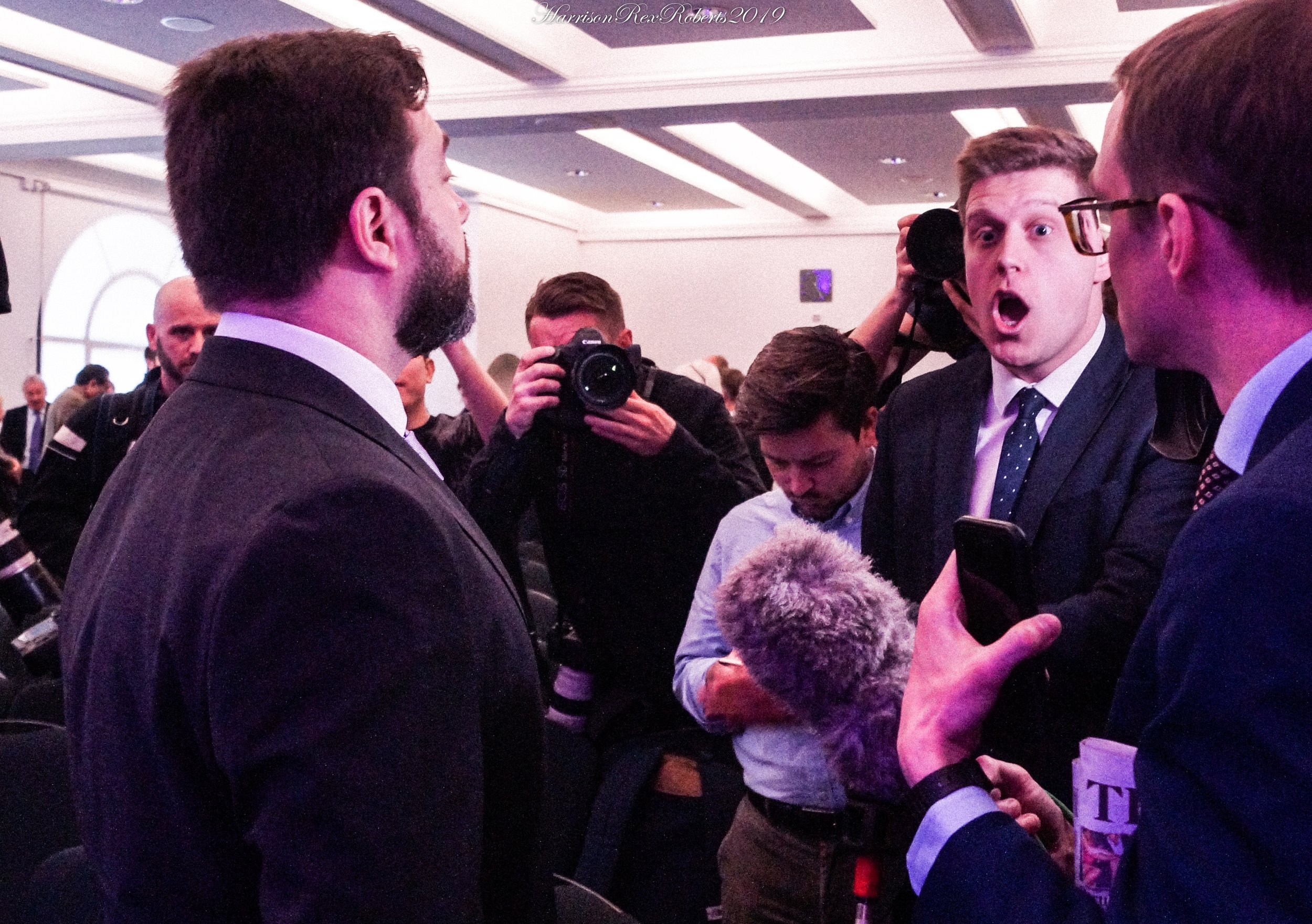 A group of men in formal attire engaged in conversation at an indoor event. One man appears surprised or expressive while the others listen or look at him. Several photographers and people areVisible in the background, indicating a busy setting.