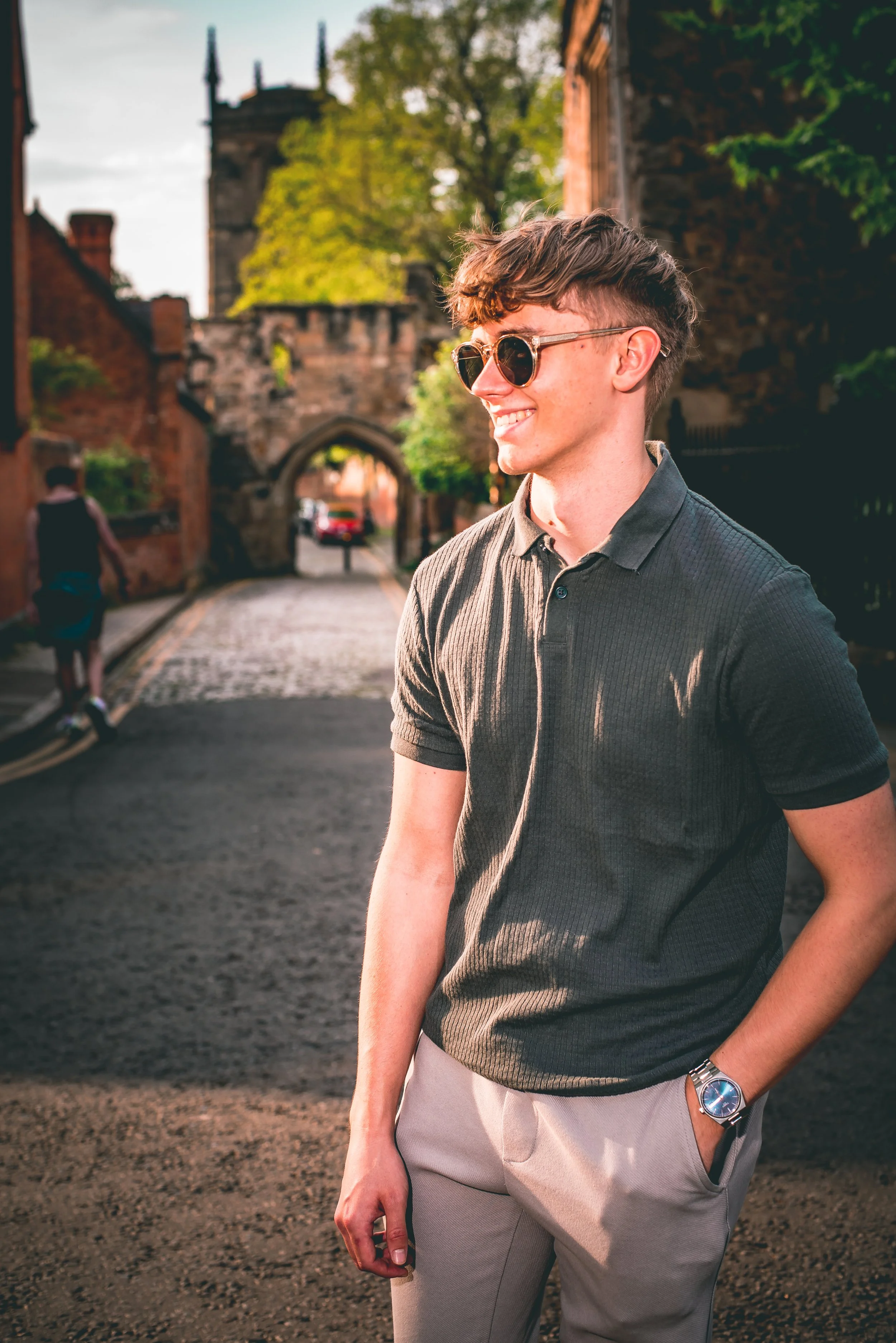 A young man with dark hair styled in a textured cut, wearing round sunglasses, a dark gray polo shirt, and beige pants, smiling and standing outdoors on a street in a historic area with brick buildings and an arched stone gateway in the background du