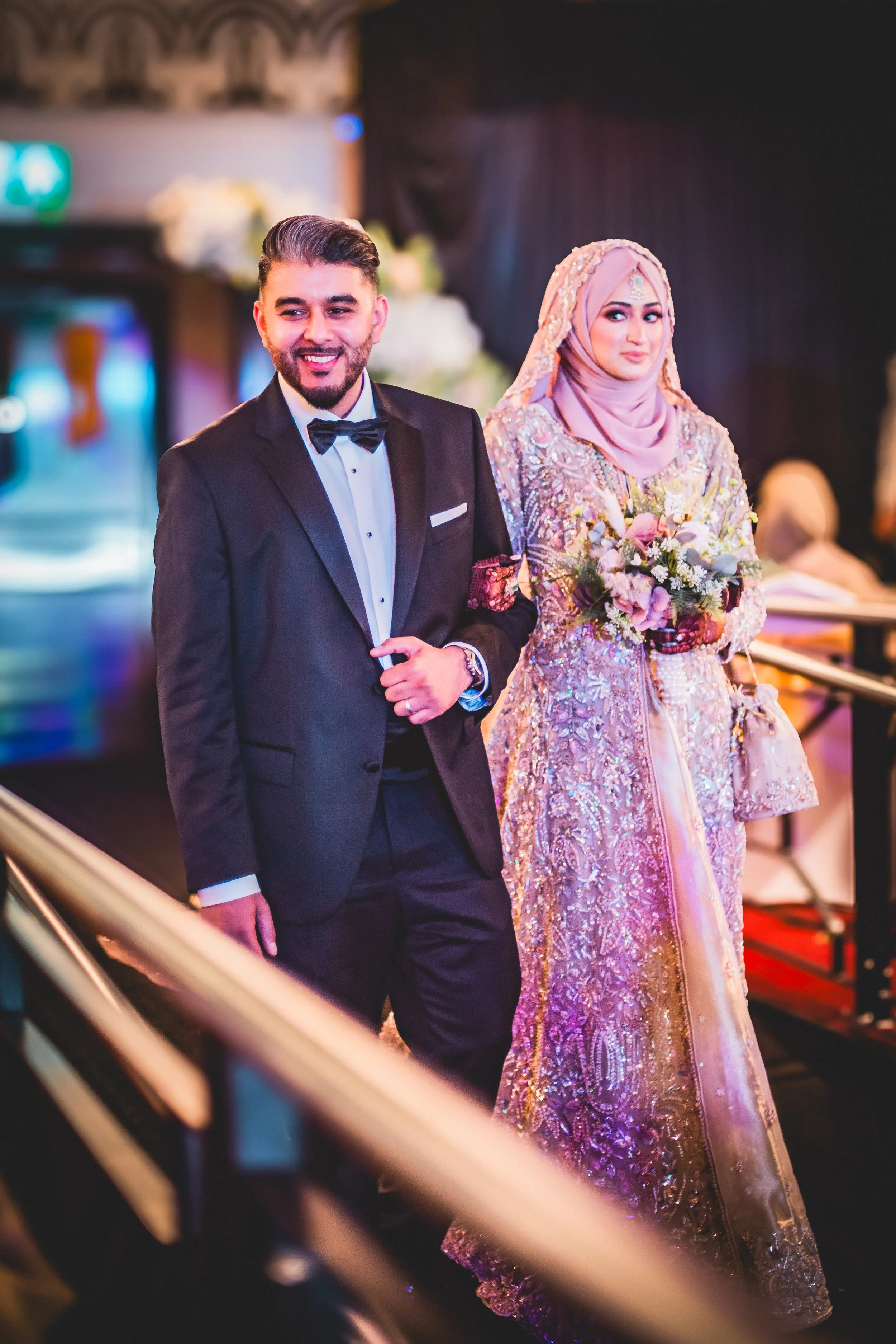 A man in a formal black tuxedo and bow tie standing next to a woman in an elaborate pink and gold traditional dress holding a bouquet of flowers at a wedding or formal event.