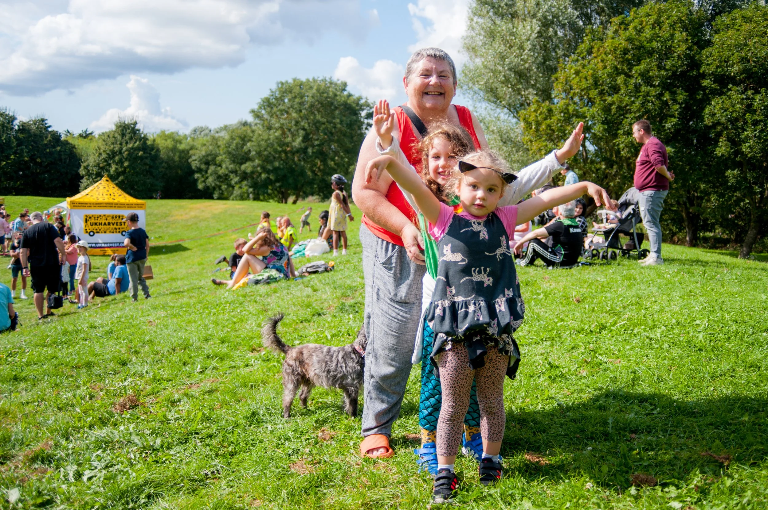A cheerful older woman and two young girls smiling and waving outdoors on a sunny day at a park or festival. The women and girls are standing on green grass, with trees and other people in the background. The older woman is wearing a red top, and the