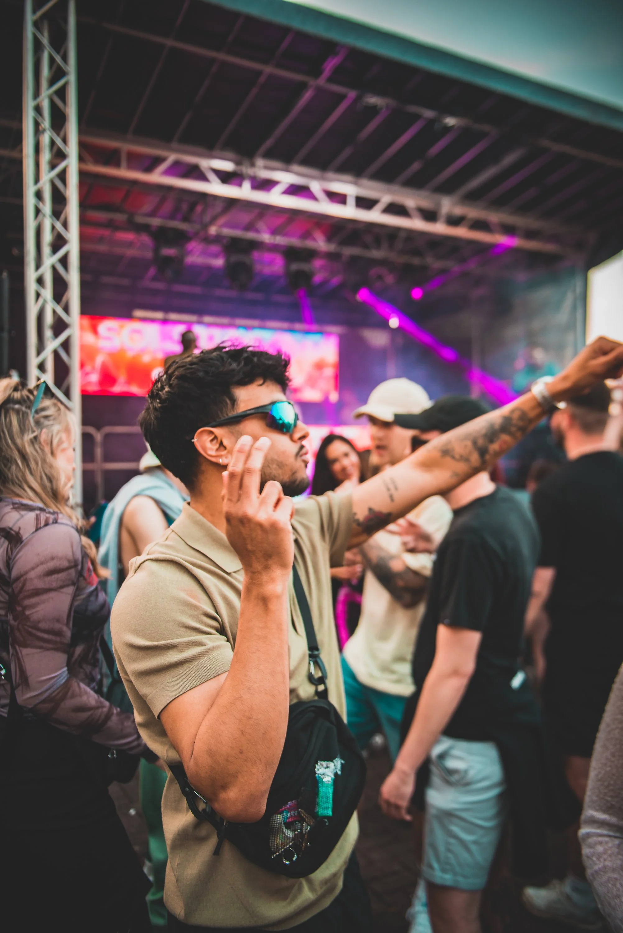 People attending an indoor concert or festival, standing close together, with a stage and colorful lighting in the background. One man is wearing sunglasses and has tattoos, holding his hand to his ear.