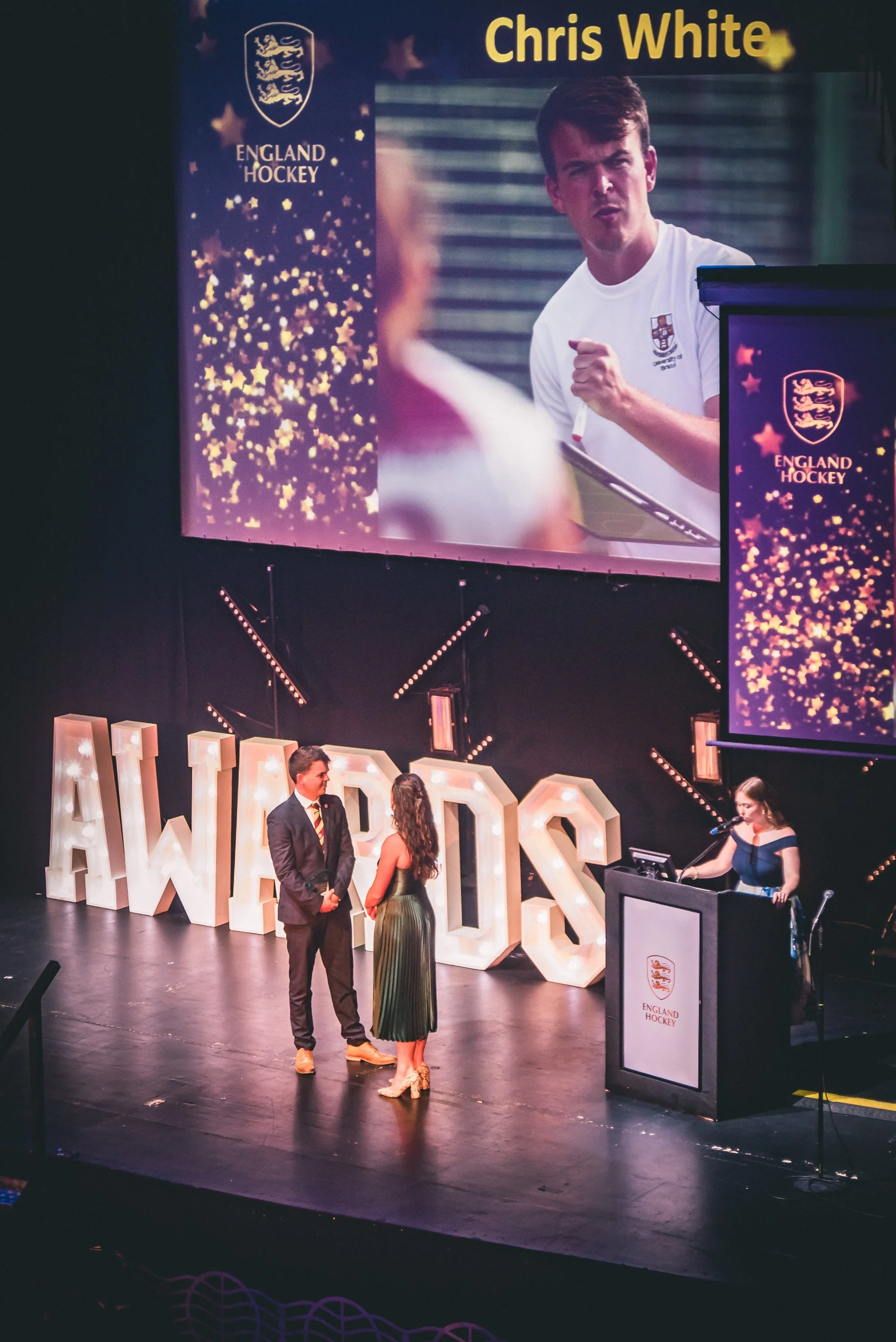 An awards ceremony or event for England Hockey, featuring large illuminated letters spelling 'AWARDS' on the stage and two people in formal attire engaging in conversation. A woman is at a podium with a microphone, and a large screen displays a pictu