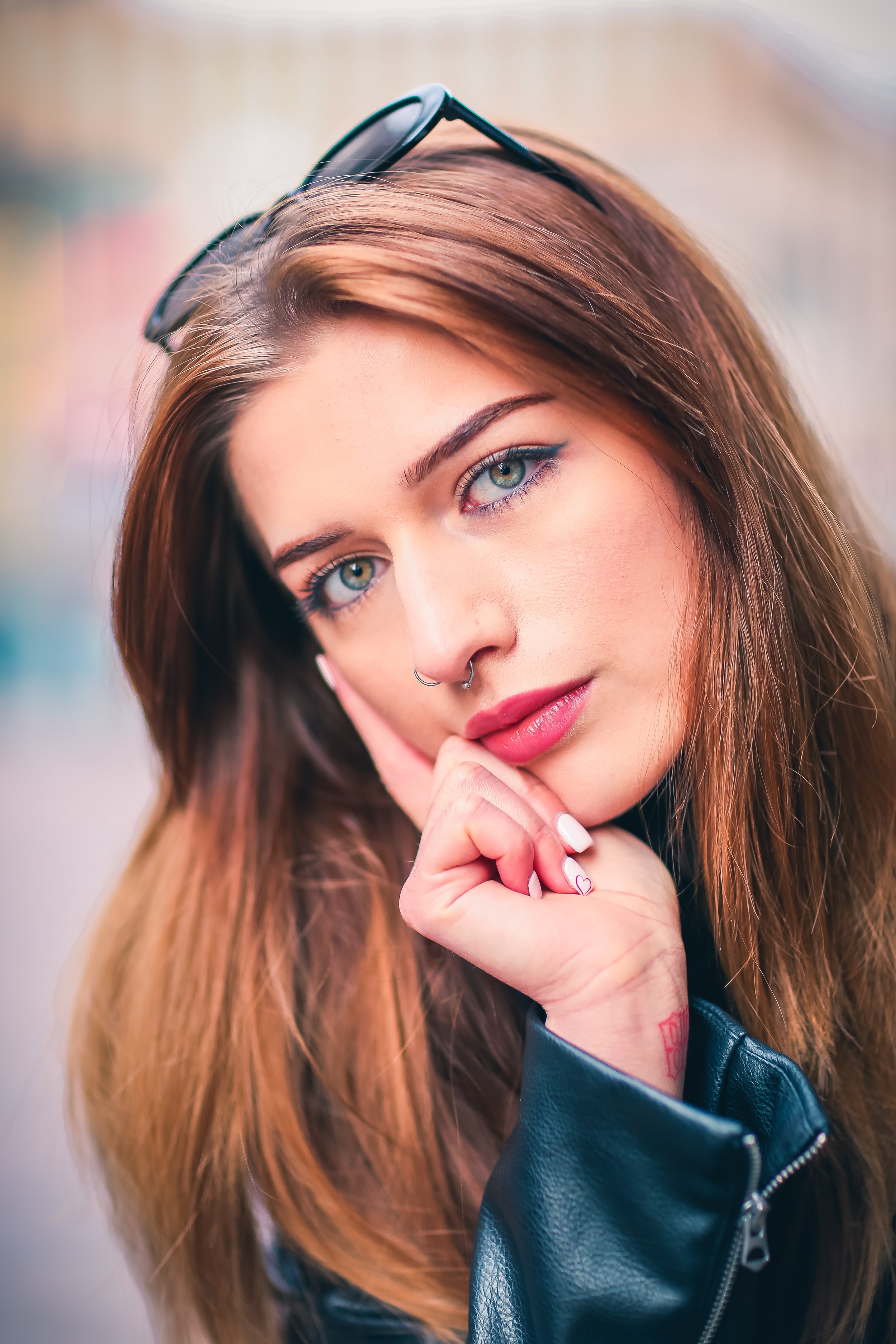 Close-up of a young woman with long, auburn hair, wearing glasses on her head, a black leather jacket, and makeup, with her hand resting near her chin.