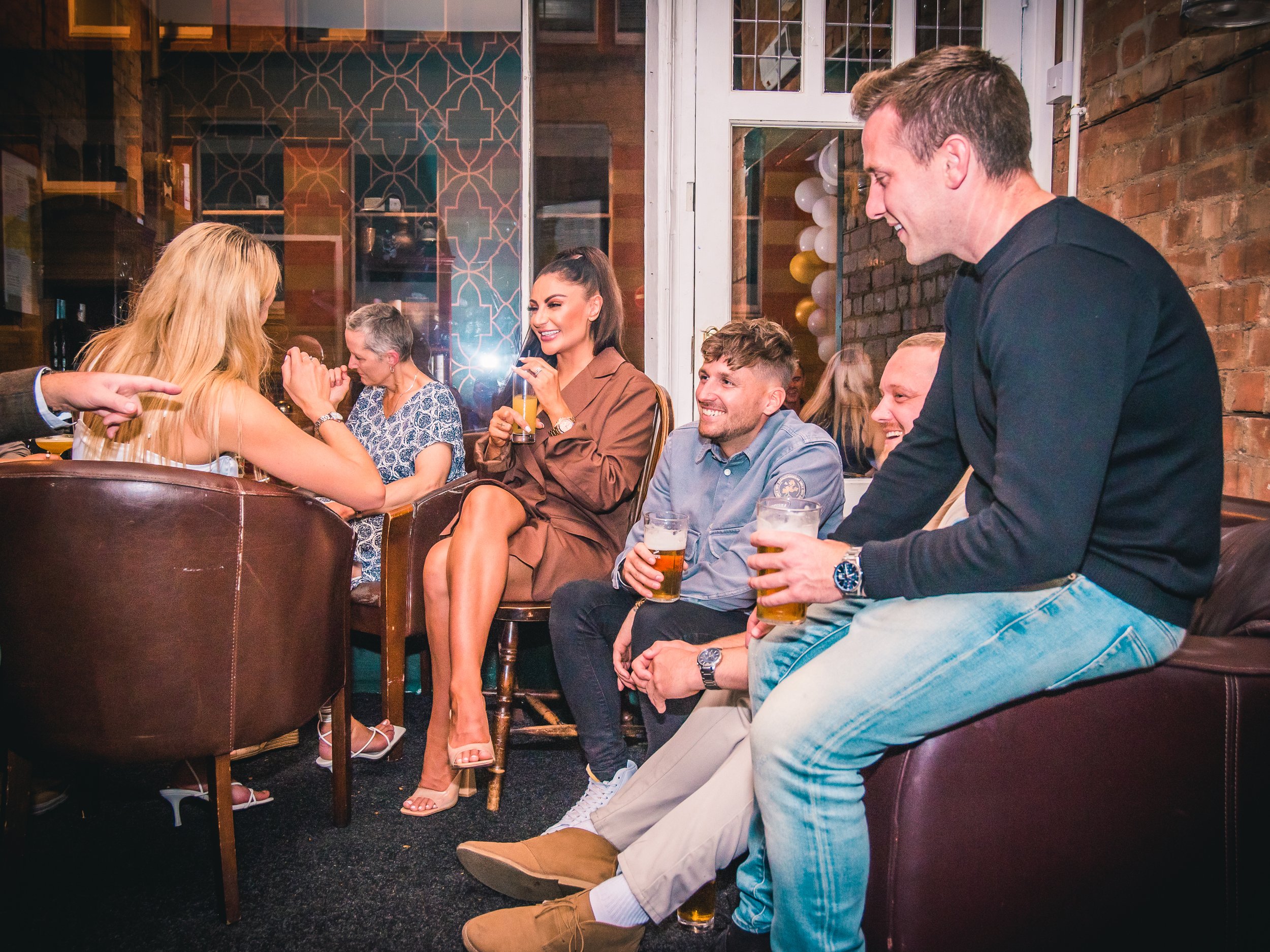 Group of friends socializing at a bar, some sitting and others standing, holding drinks, in a cozy, dimly lit indoor setting with brick walls and decorative wallpaper.