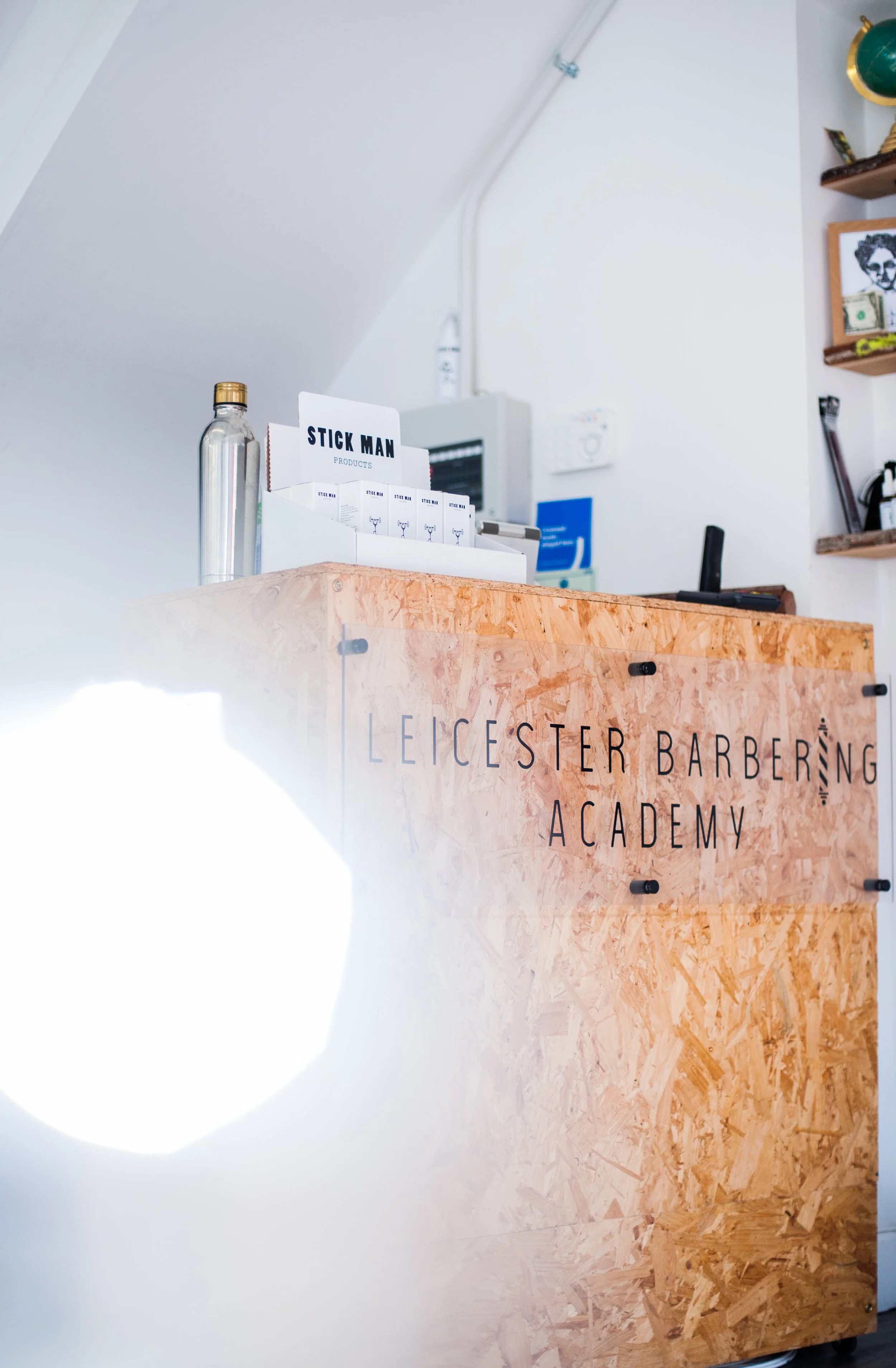 Wooden reception desk with the words 'Leicester Barbering Academy' printed on it, a clear water bottle, and some leaflets or product samples. In the background, a white wall, shelves with various items, and a control panel are visible.