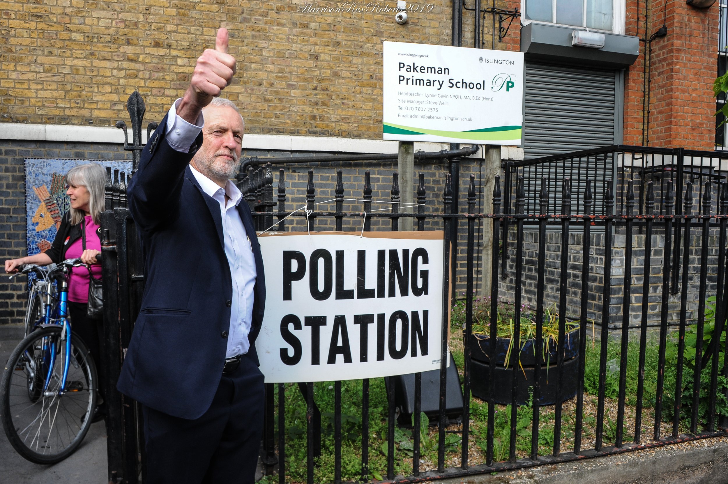 A man with gray hair and a beard giving a thumbs-up gesture in front of a school building with a sign that reads "Pakeman Primary School". A woman with gray hair and a pink jacket is pushing a bicycle nearby. The scene is outdoors, near a black iron 