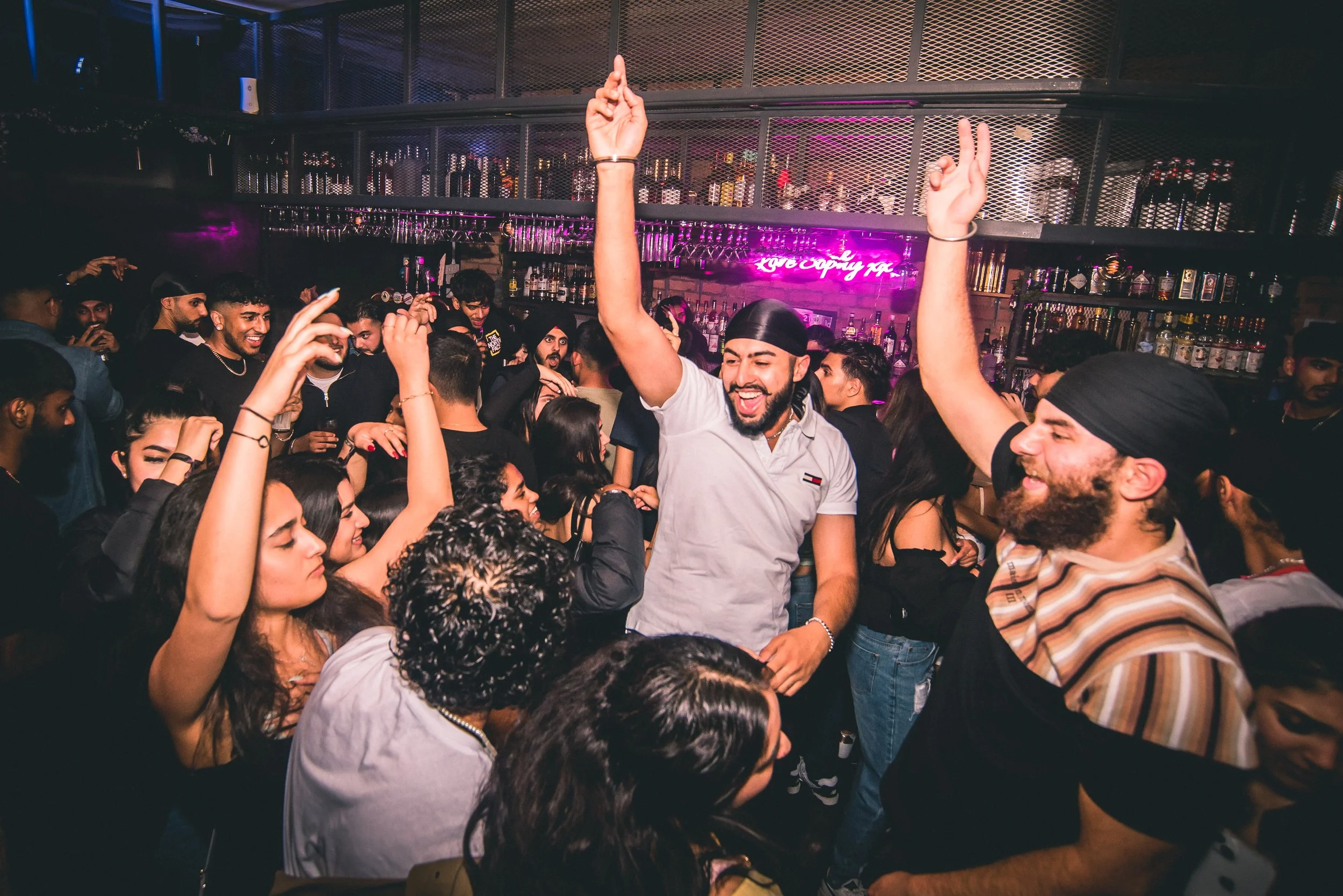 A lively nightclub scene with people dancing and socializing, illuminated by purple and pink neon lights, with a bar stocked with bottles in the background.