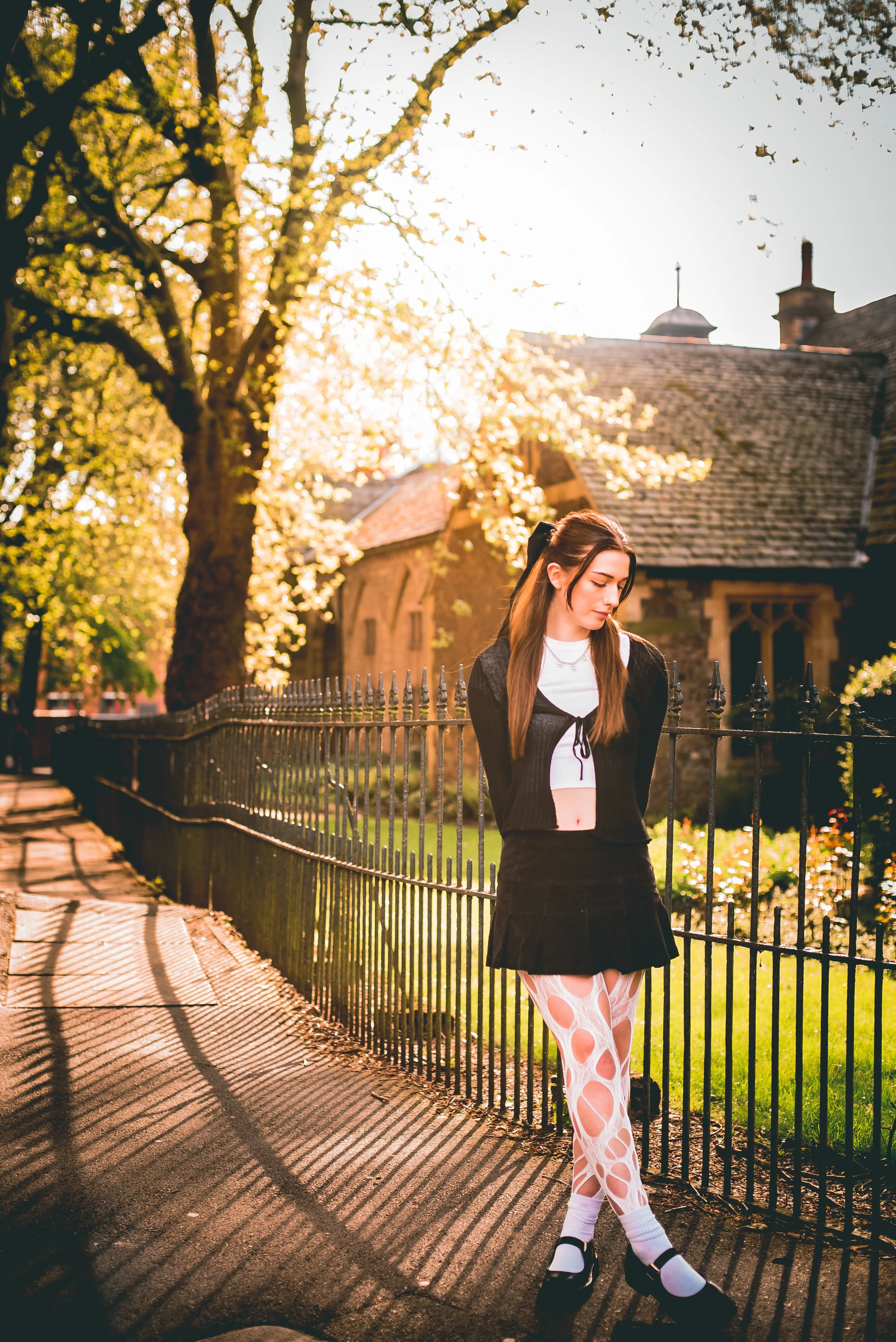 A young woman with long brown hair standing outdoors near a black metal fence with a shadow pattern on the sidewalk, wearing a black jacket, white top, and fashionable ripped patterned tights, with an old building and trees in the background during t