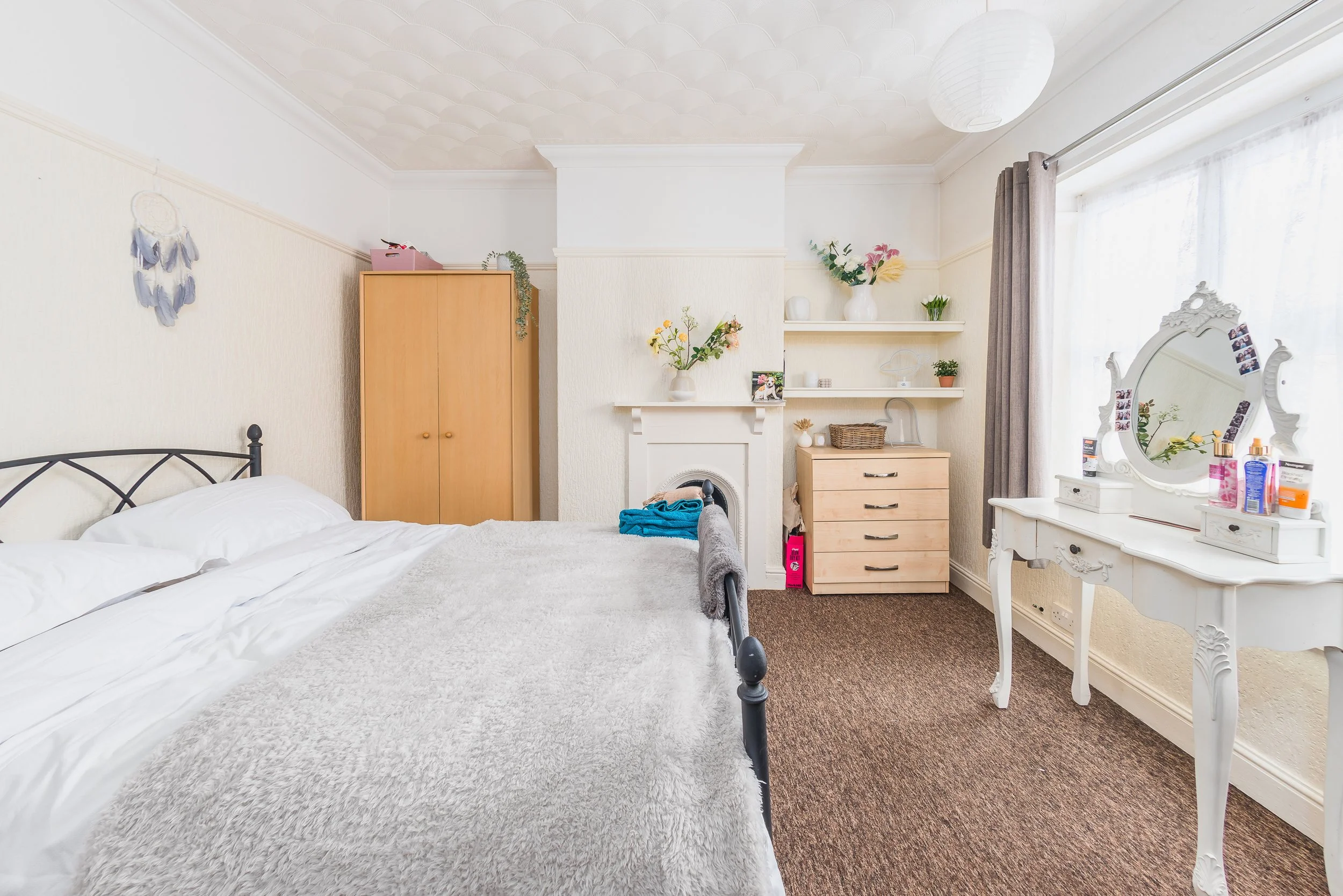 A bedroom with two beds with white bedding, a wooden wardrobe, a white fireplace mantel with floral arrangements, a set of white shelves with decorative items, and a vintage white dresser with a mirror and photo clips, near a window with gray curtain