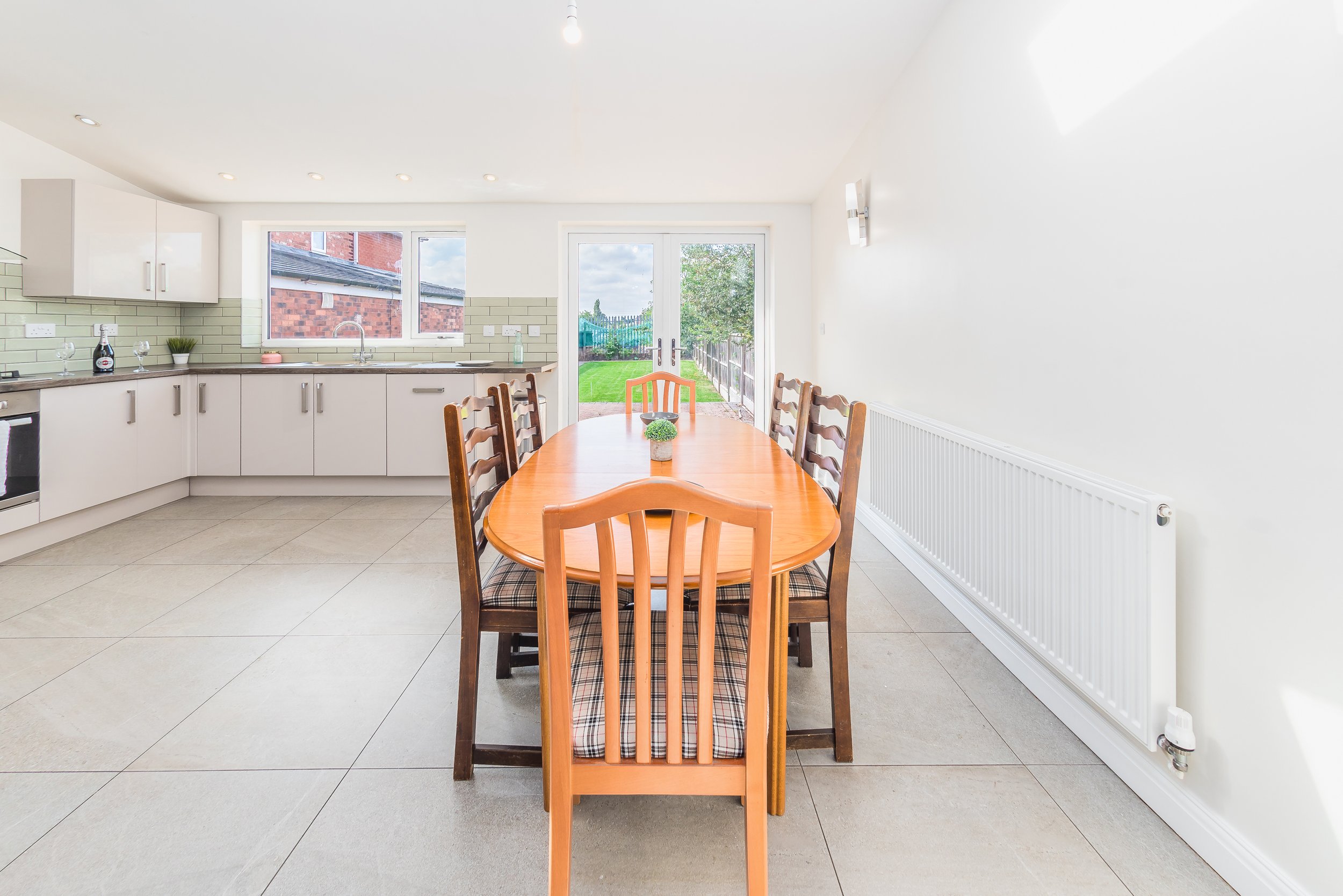 A dining area with a wooden table and six chairs, white kitchen cabinets, a window, a glass door leading to a backyard, and a radiator on the right wall.