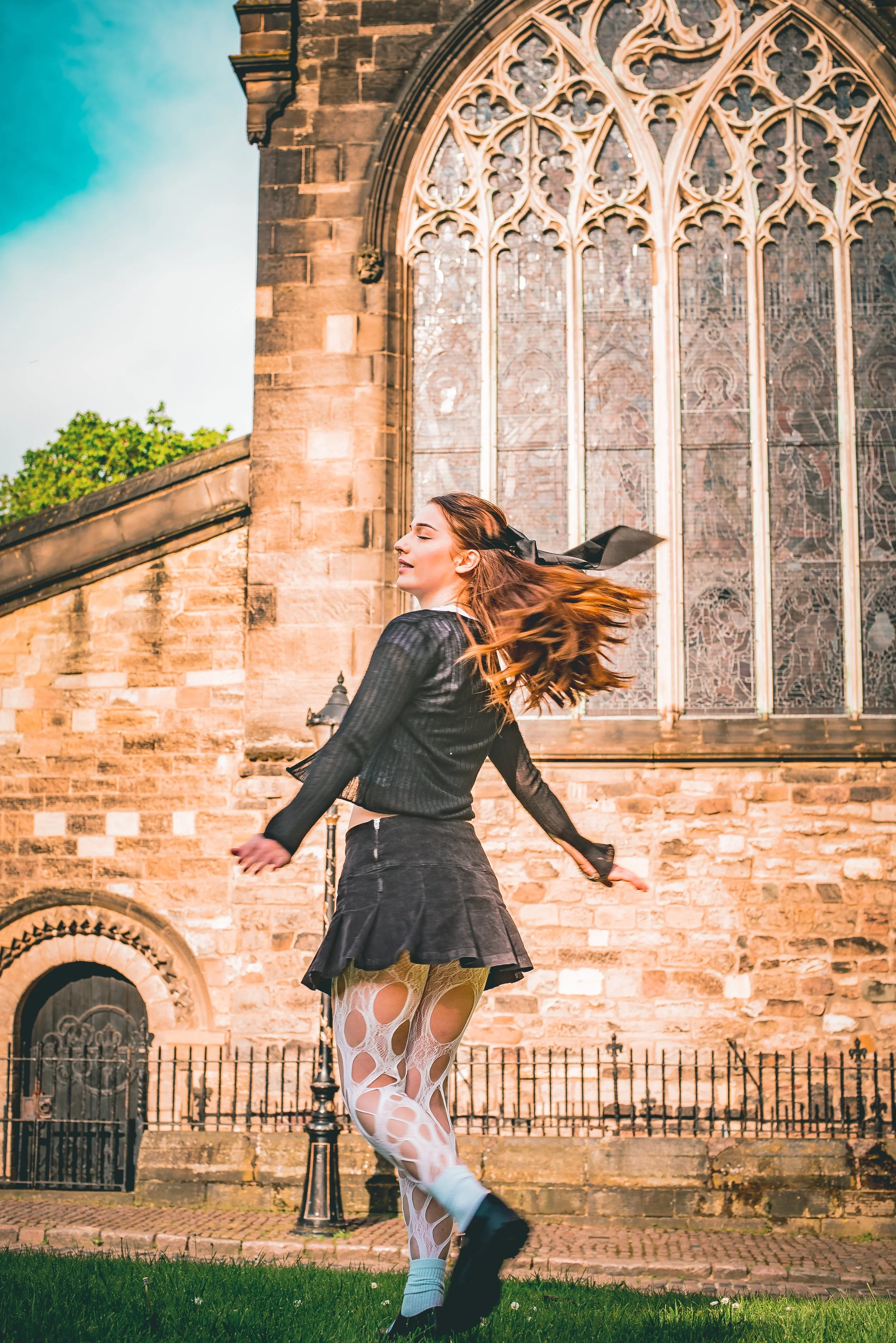 Young woman with long hair dancing or spinning in front of a historic stone church with stained glass windows, wearing a black jacket, skirt, and white leggings with large circular cutouts, outdoors during daytime.
