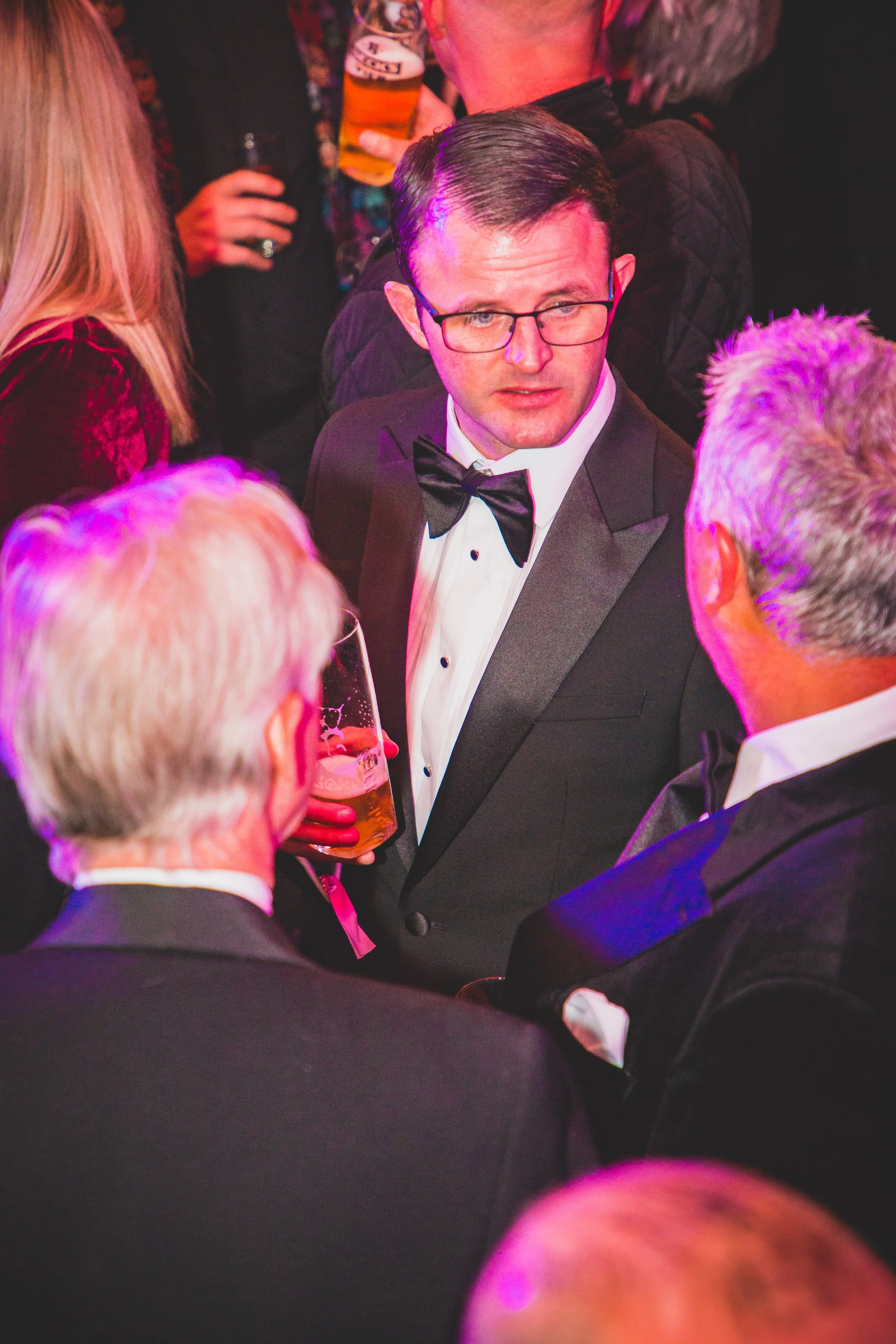 A man wearing glasses, a tuxedo, and a bow tie at a social event, holding a glass of beer and engaging in conversation with others.