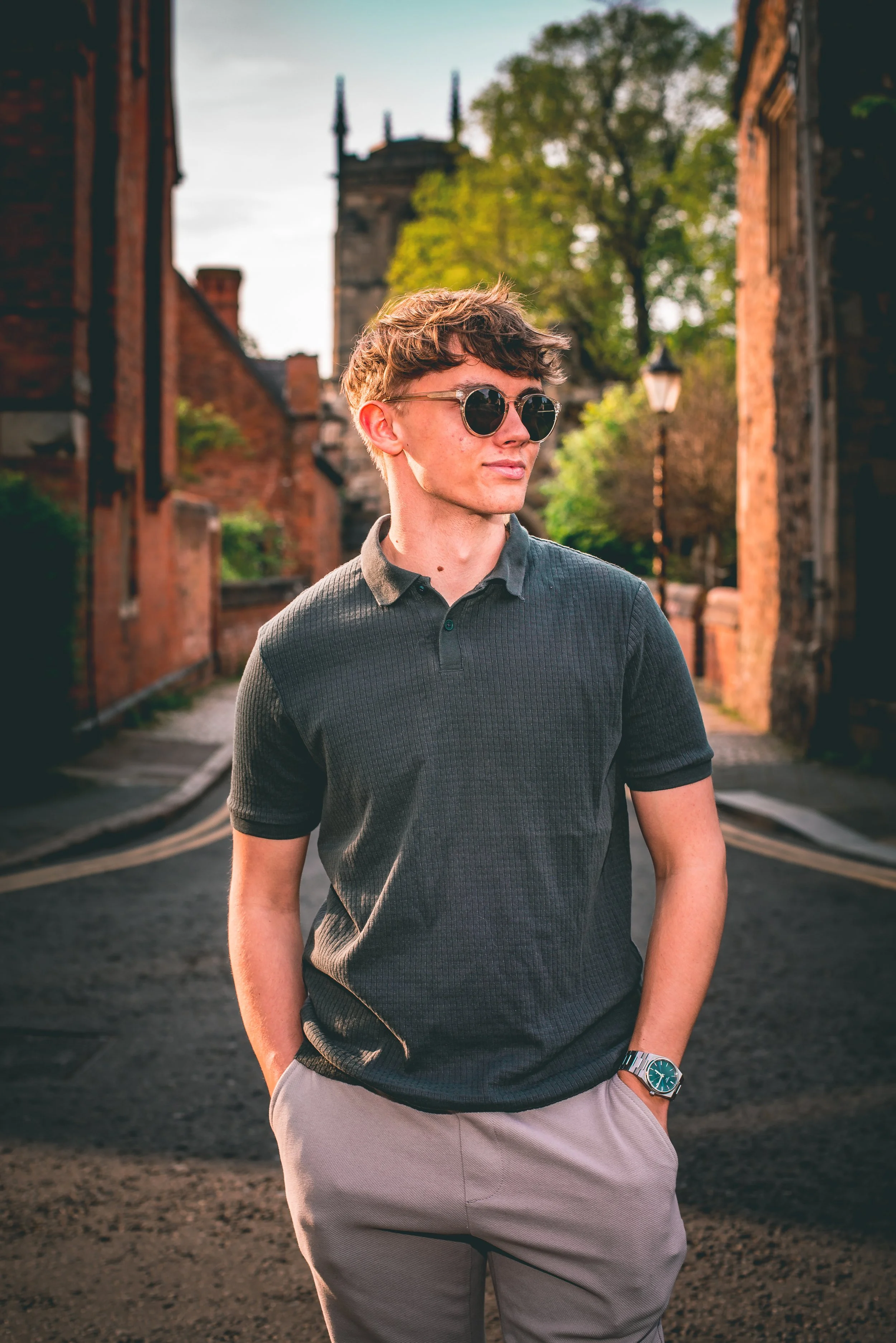 A young man with wavy hair wearing sunglasses, a dark short-sleeved polo shirt, light-colored pants, and a wristwatch, walking outdoors on a street lined with old brick buildings and a green tree in the background.