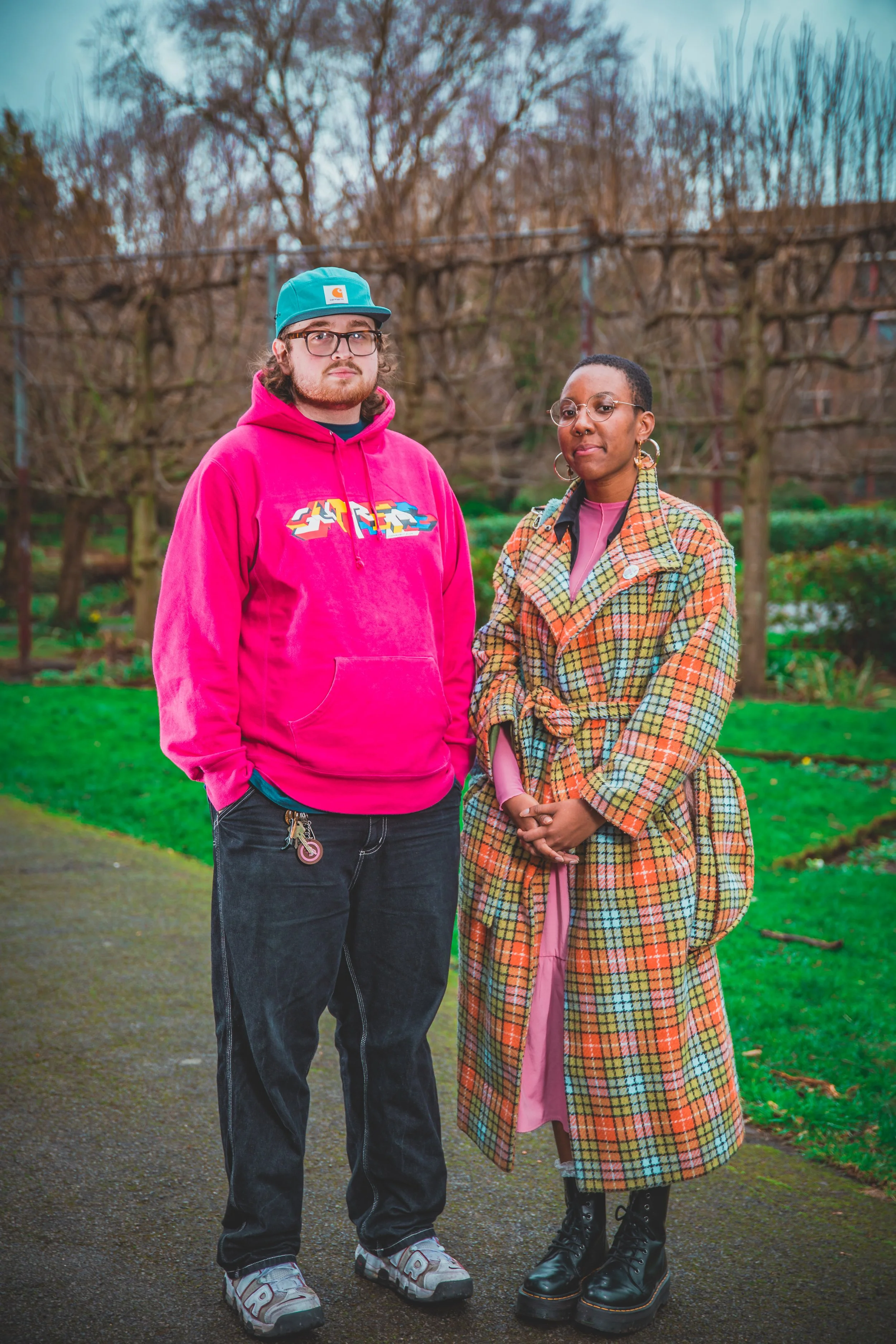 A man and a woman stand outdoors on a paved path in a park or garden with trees and greenery in the background. The man is wearing a bright pink hoodie with a graphic design, black jeans, gray sneakers, and a teal cap with a logo, and has glasses and
