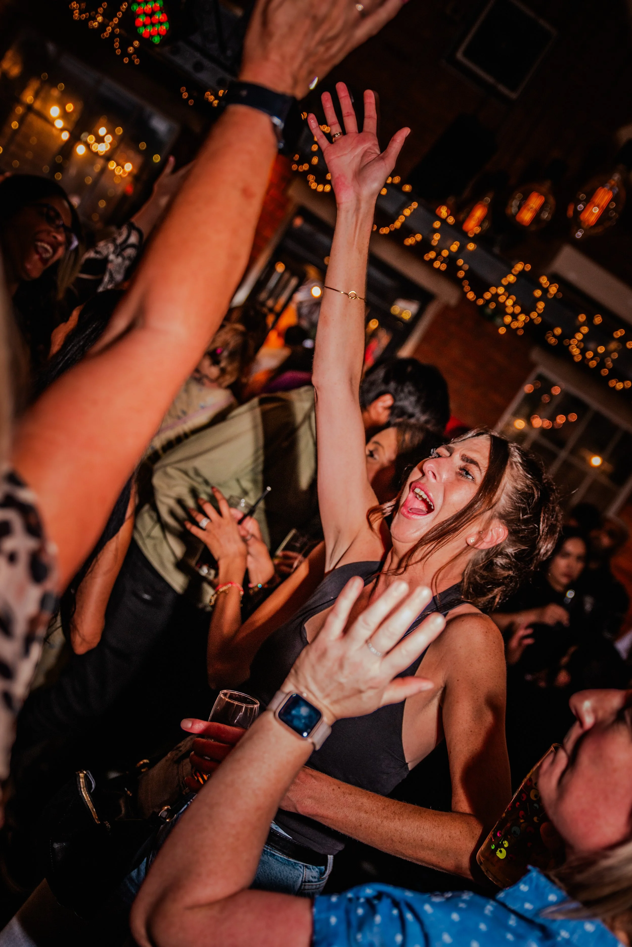 A group of people dancing and celebrating inside a dimly lit venue with warm lighting and string lights hanging from the ceiling.