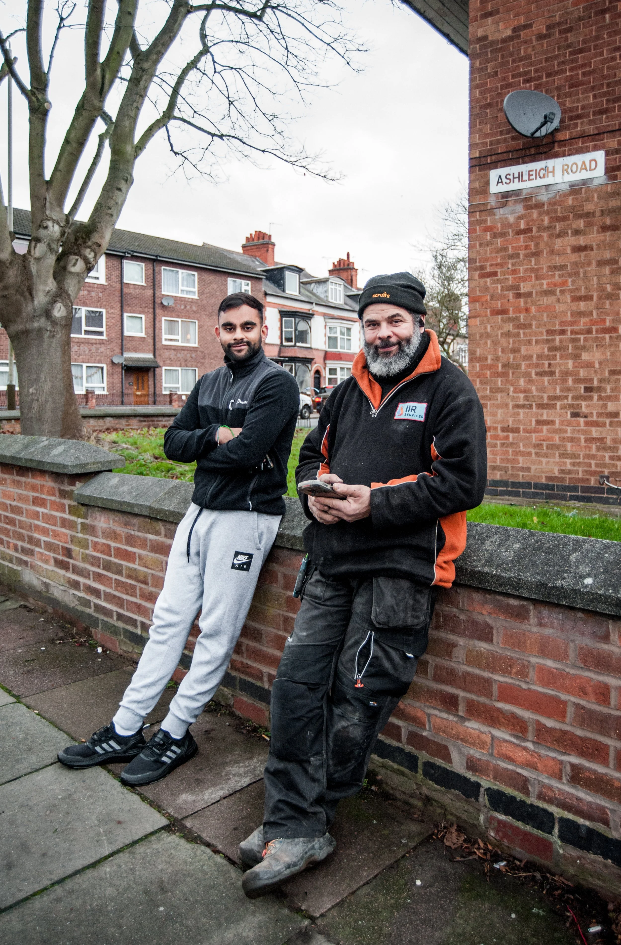 Two men standing against a brick wall on Ashleigh Road, one wearing a black jacket and gray sweatpants, the other in a black and orange jacket and black pants, both smiling, standing with arms crossed or holding a phone, with residential buildings in
