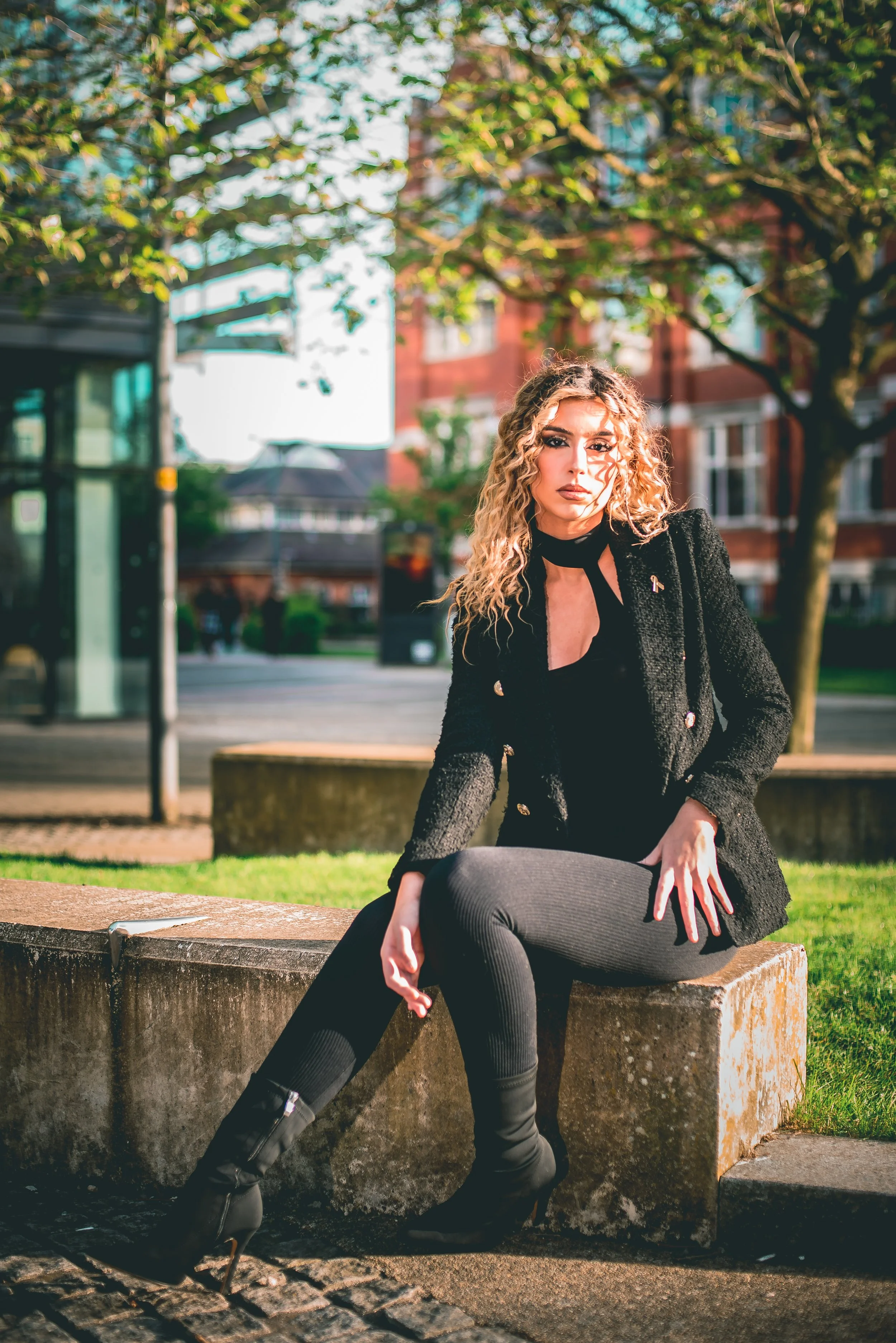 A woman with curly blonde hair and dark makeup sitting on a stone bench outdoors, dressed in black leather pants, a black top, and a textured black blazer, with a background of trees and buildings.