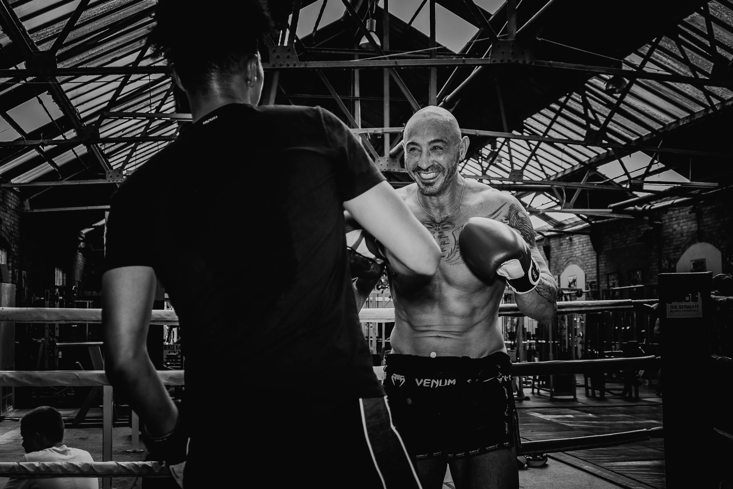 A shirtless male boxer with tattoos on his chest and arms wearing boxing gloves, smiling and standing in a gym under a metal roof, as another person in black workout clothes faces him.