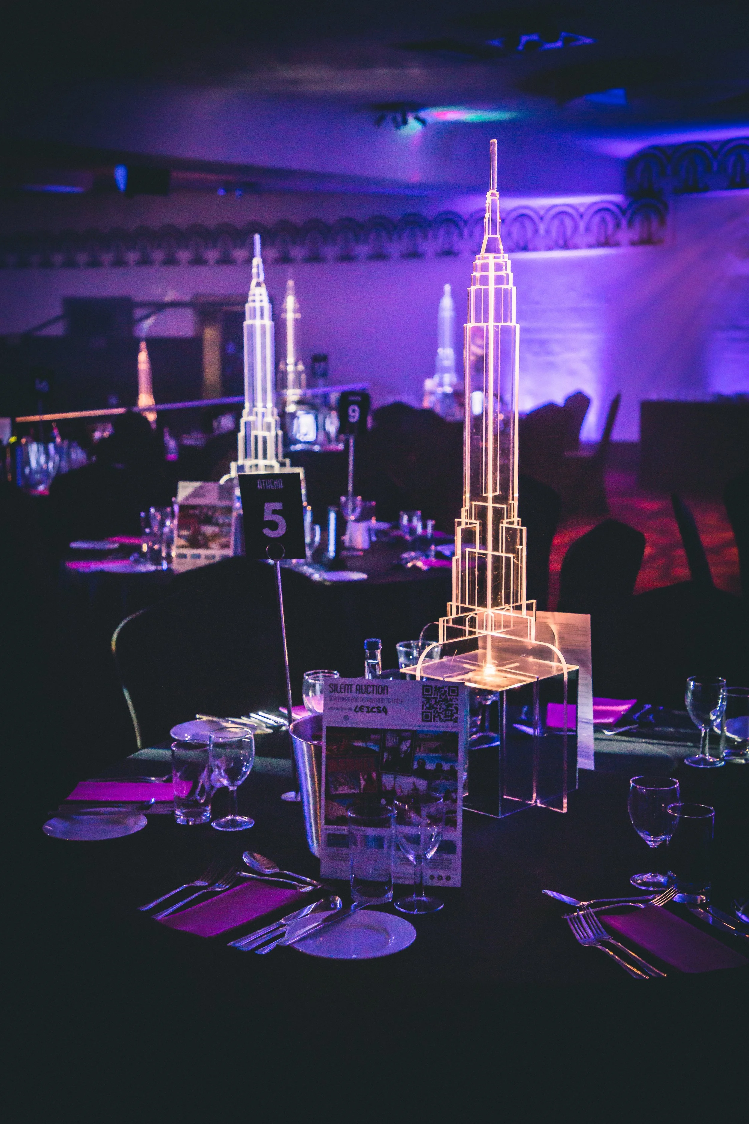 LED table centerpiece resembling a skyscraper at a formal event with tables, napkins, glasses, and silverware in a dimly lit room with purple lighting.