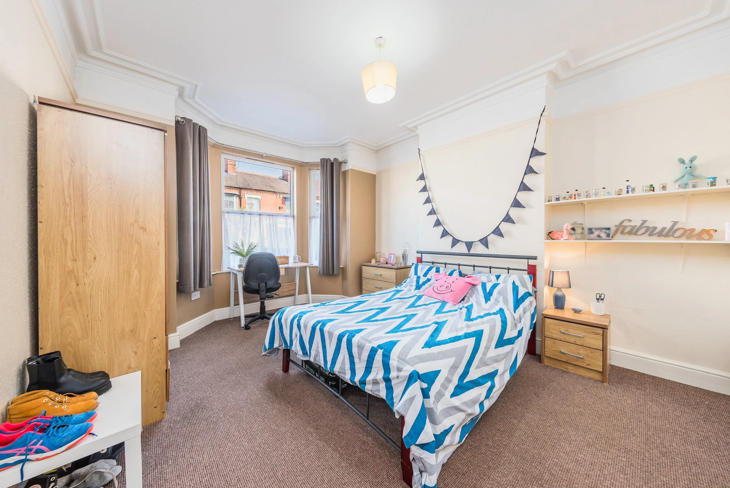 Bedroom with a bed covered in a blue and white zigzag pattern comforter, a pink Peppa Pig pillow, two wooden nightstands, a shelf with toys and decorations, a window with curtains, a desk with a chair, and shoes on the floor.