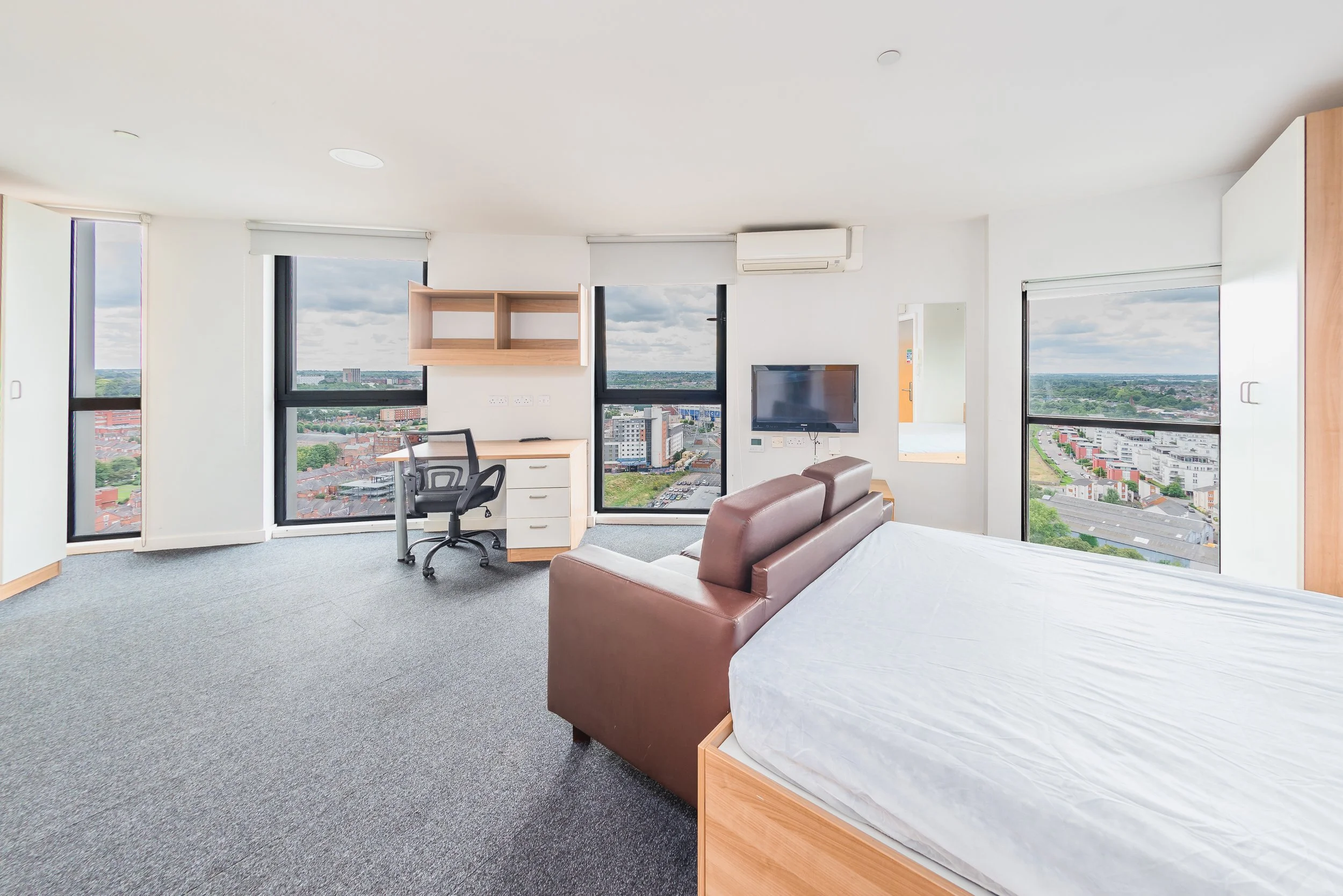 Interior of a high-rise apartment with large windows overlooking a cityscape, furnished with a desk, chair, brown sofa, white bed, and a wall-mounted TV.