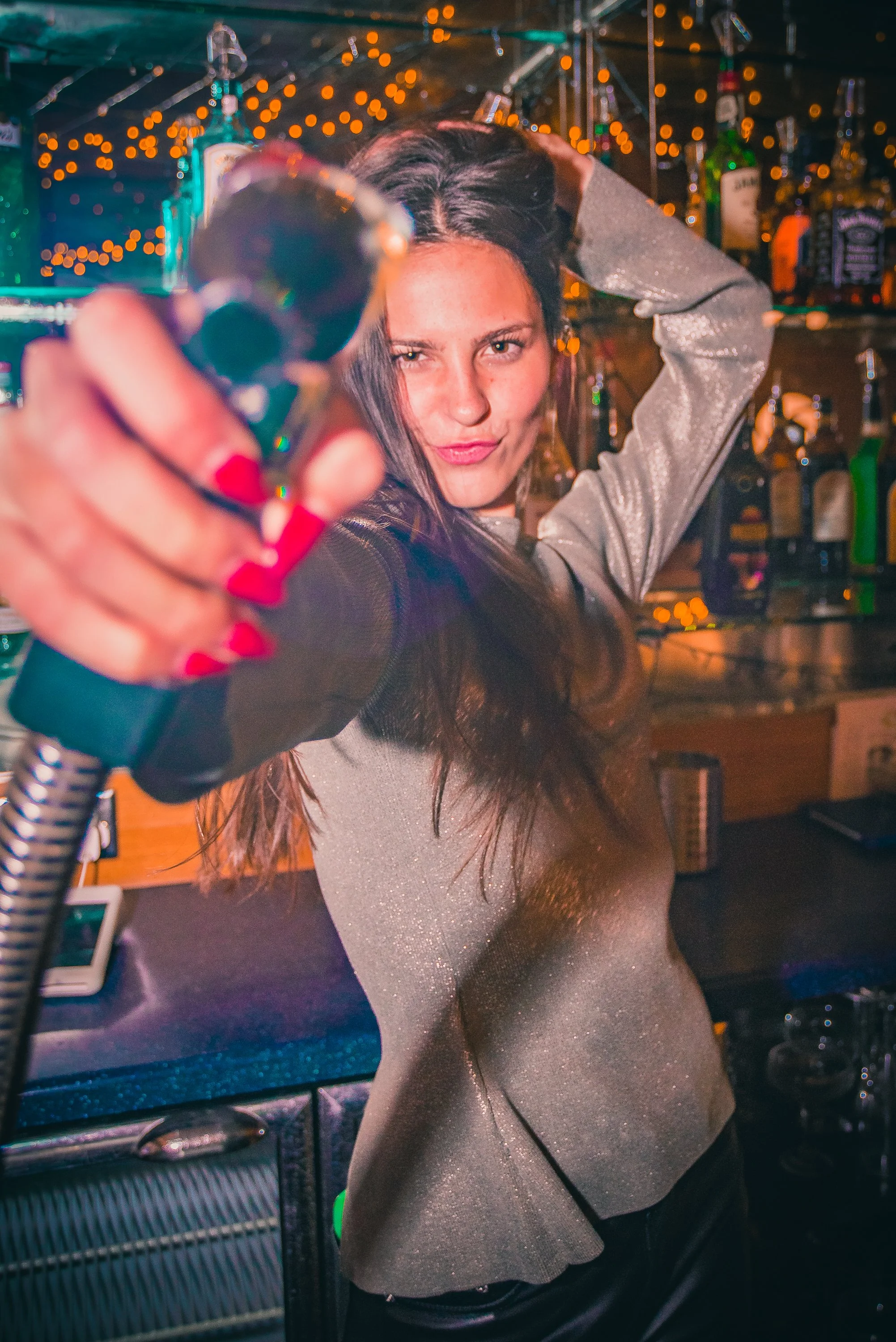 A woman with long dark hair is holding a cocktail glass towards the camera, wearing a shiny, silver top. She appears to be in a bar with colorful bottles and string lights in the background.