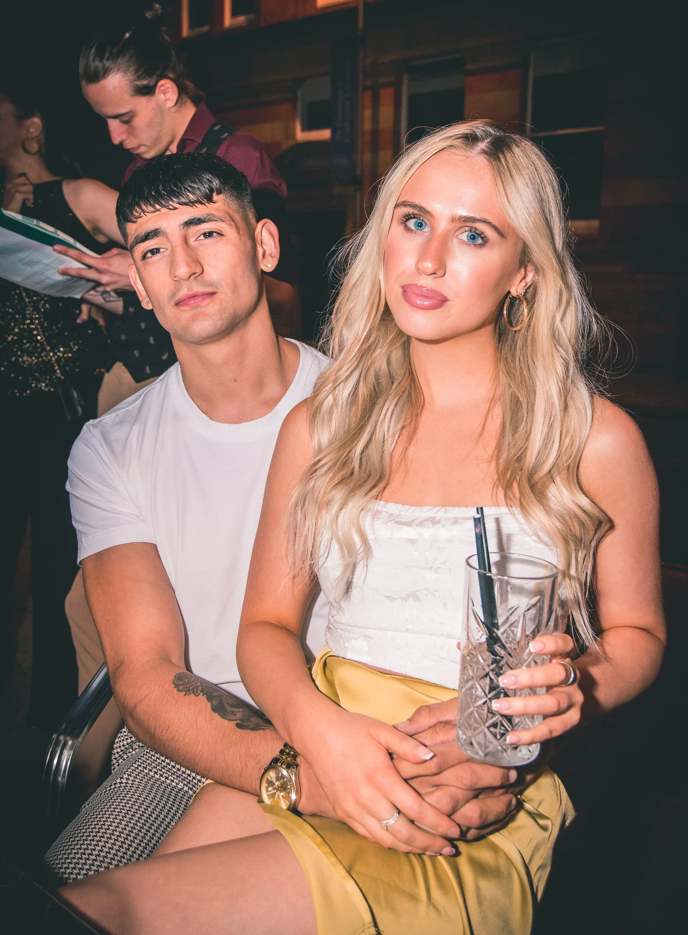 A young man and woman sitting closely at a night event, the woman holding a glass with a black straw. The woman has long blonde hair, blue eyes, and is wearing gold hoop earrings and a white top with yellow shorts. The man has short dark hair, a tatt