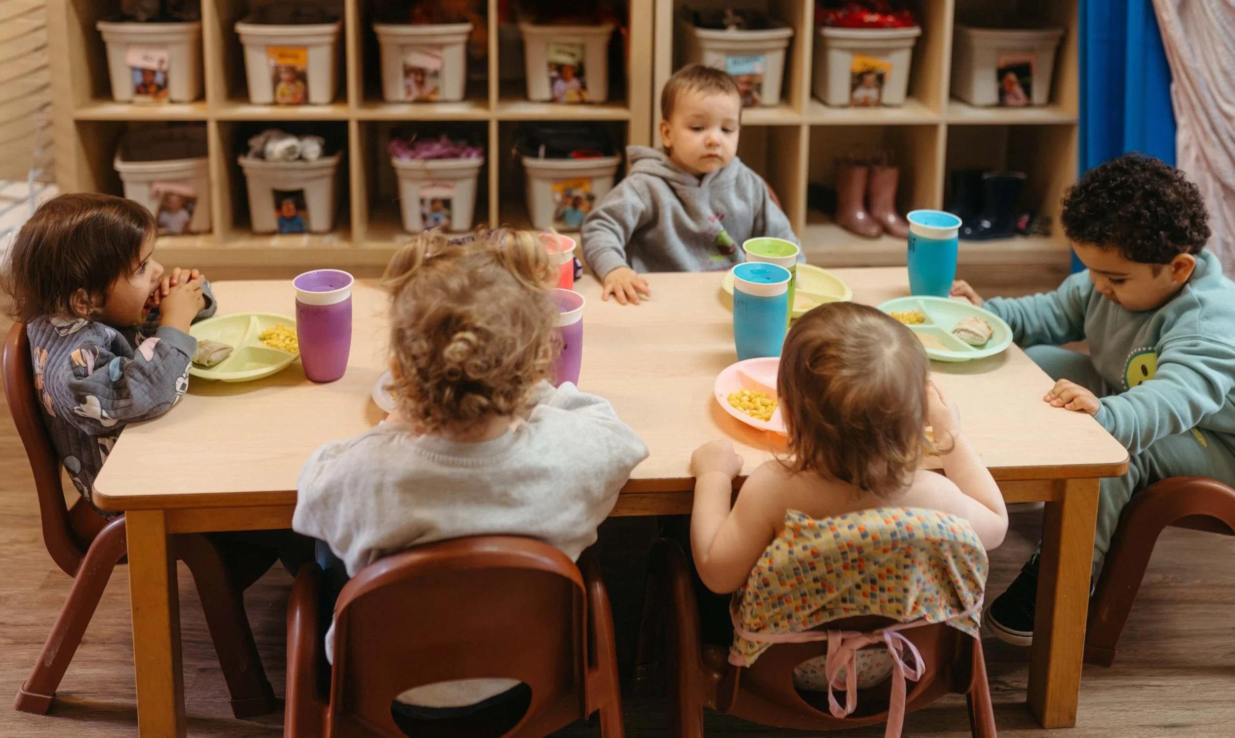 Children sitting at a table with colorful trays and cups, in a classroom with storage bins in the background.