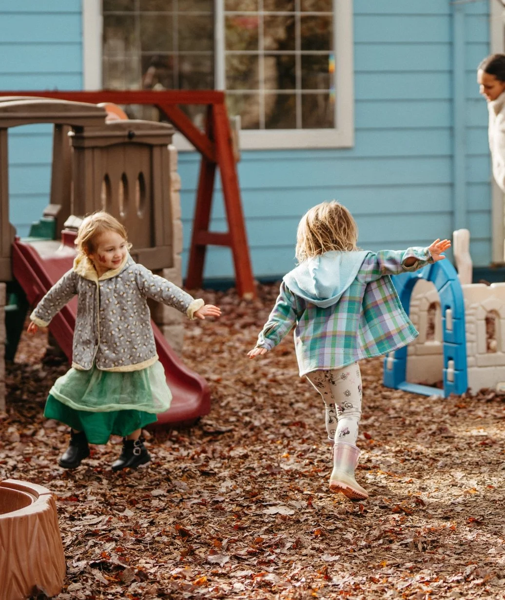Two young children playing and running on a leaf-covered playground in autumn, near a blue house with a window, with a woman watching in the background.