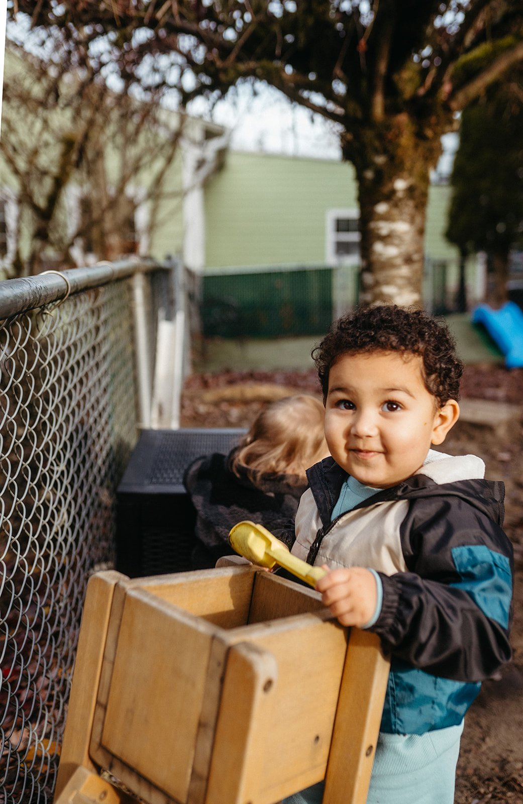 Young boy with curly hair holding a yellow toy shovel and a wooden crate outdoors.