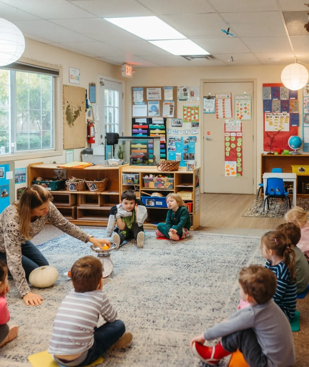 A classroom with children seated on a carpet, participating in a teaching activity with a teacher. The classroom contains educational decorations, shelves with supplies, and colorful posters on the walls.
