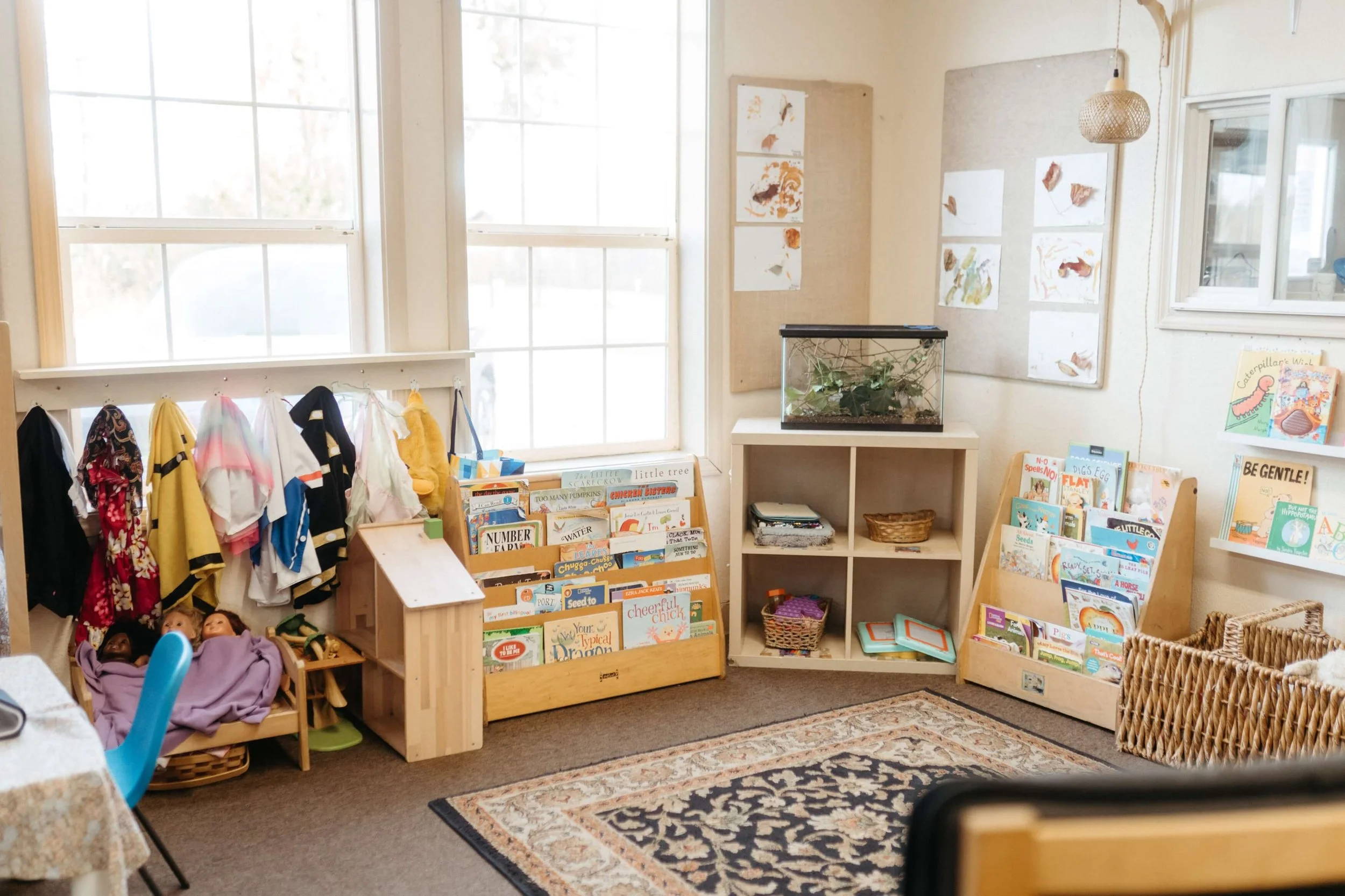 A classroom corner with shelves of children's books, a small fish tank, children's clothing hanging, and artwork on the walls, illuminated by natural light from large windows.