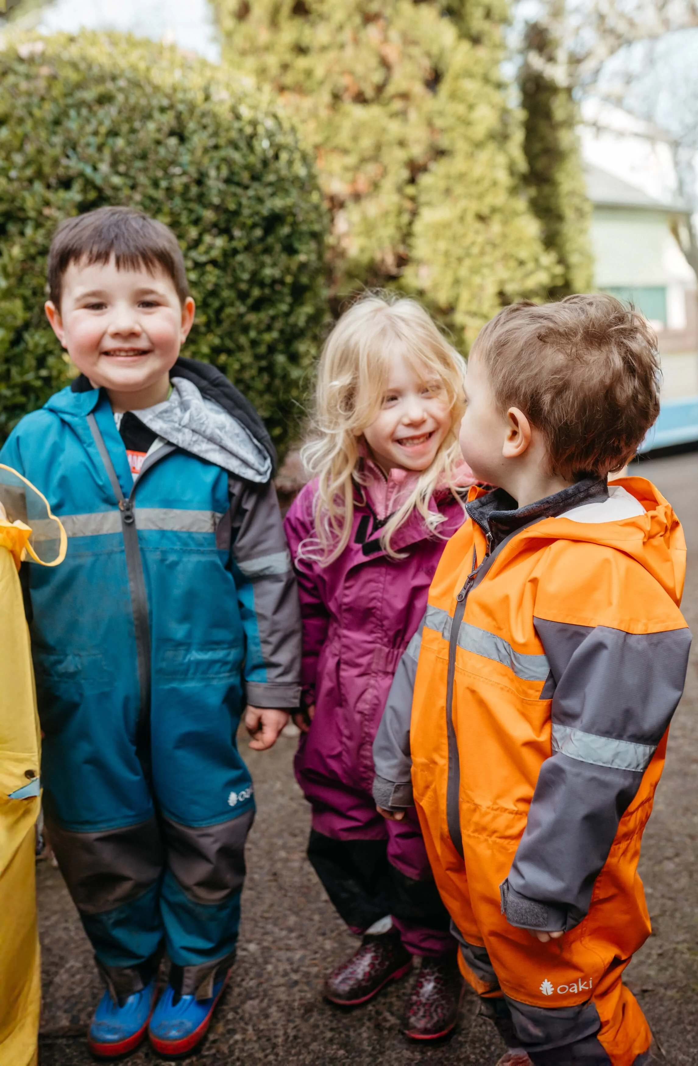 Three children outdoors in jackets, standing close together, smiling and talking to each other with trees and a house in the background.