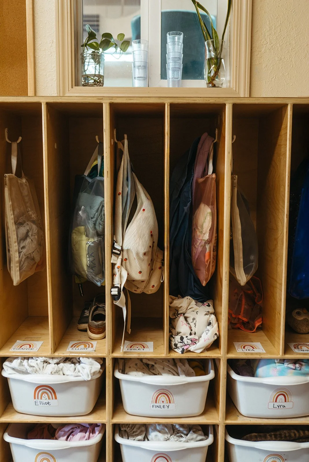 Wooden cubbies filled with backpacks and plastic bins labeled with names, set against a wall with a mirror and vases of plants above.