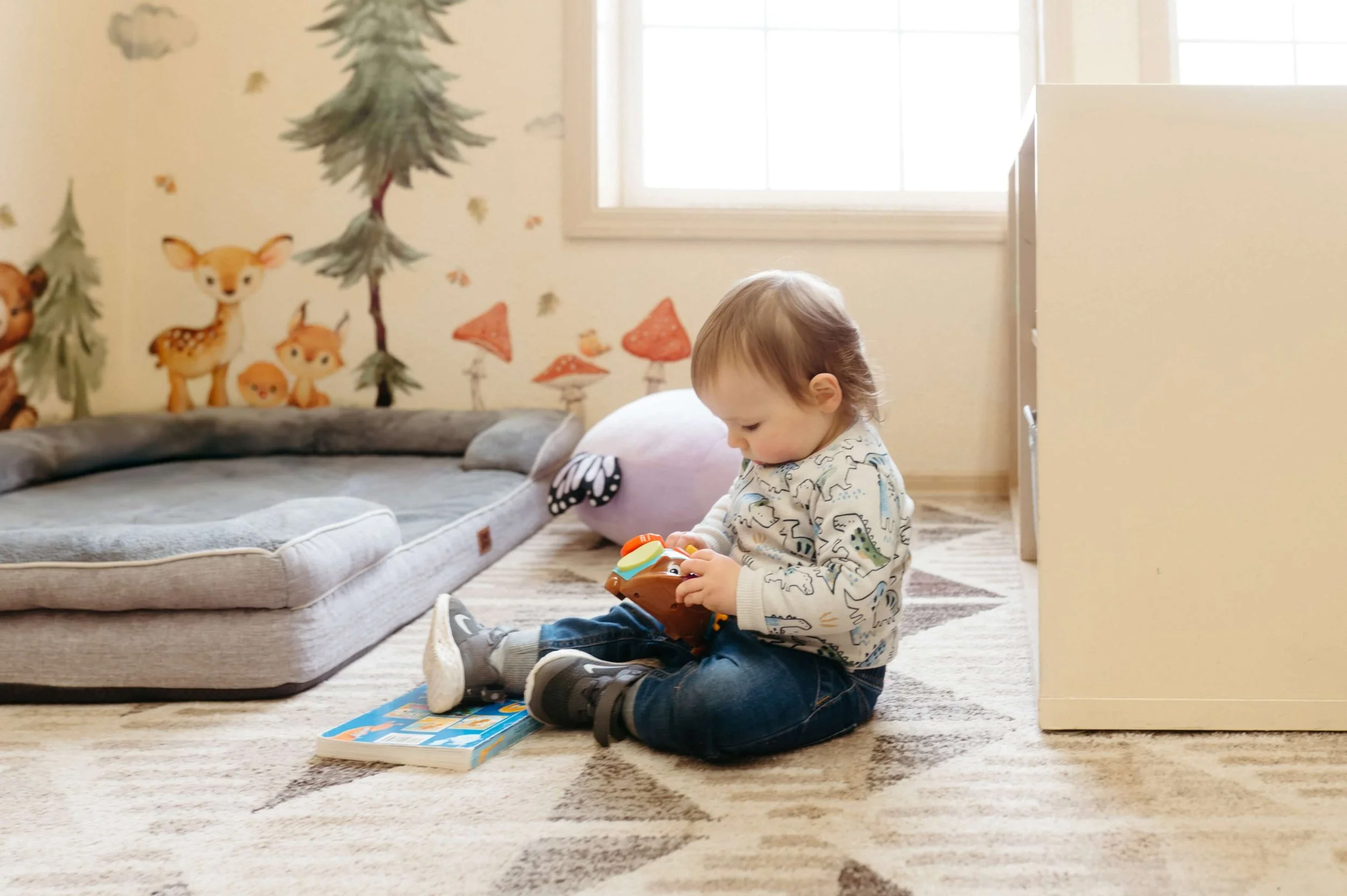 A young boy sitting on a carpeted floor playing with a toy in a brightly lit room with a gray sofa, a plush pillow, and a colorful wall mural of animals and trees.