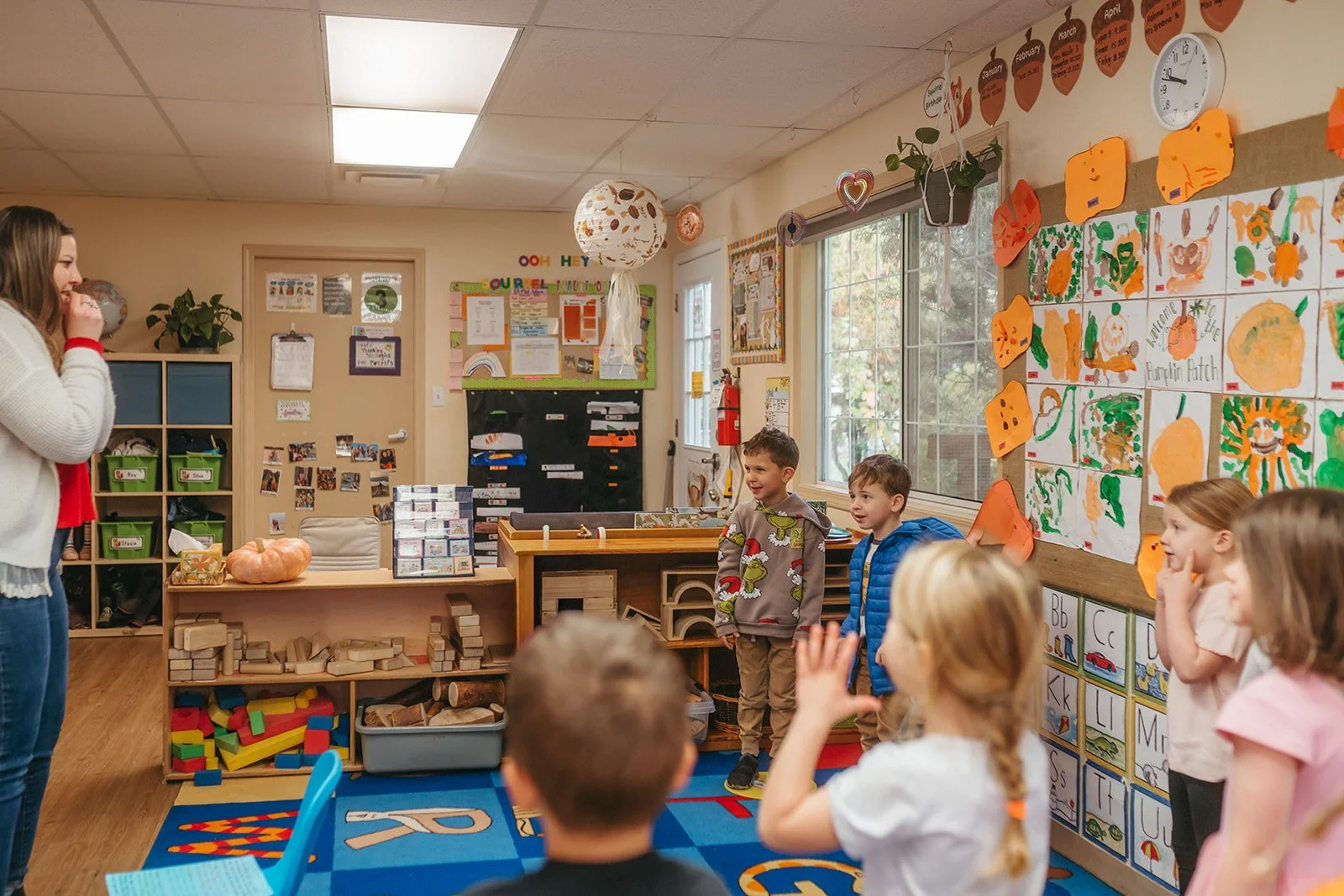 A kindergarten classroom with children standing and a teacher on the left. The room has colorful decorations, artwork, and educational materials, with wooden blocks and toys on shelves.