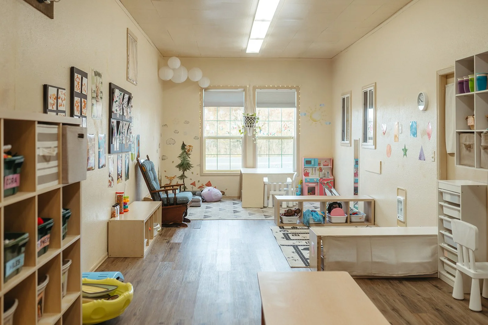 A children's playroom with shelves, toys, and artwork on the walls, illuminated by natural light from large windows.