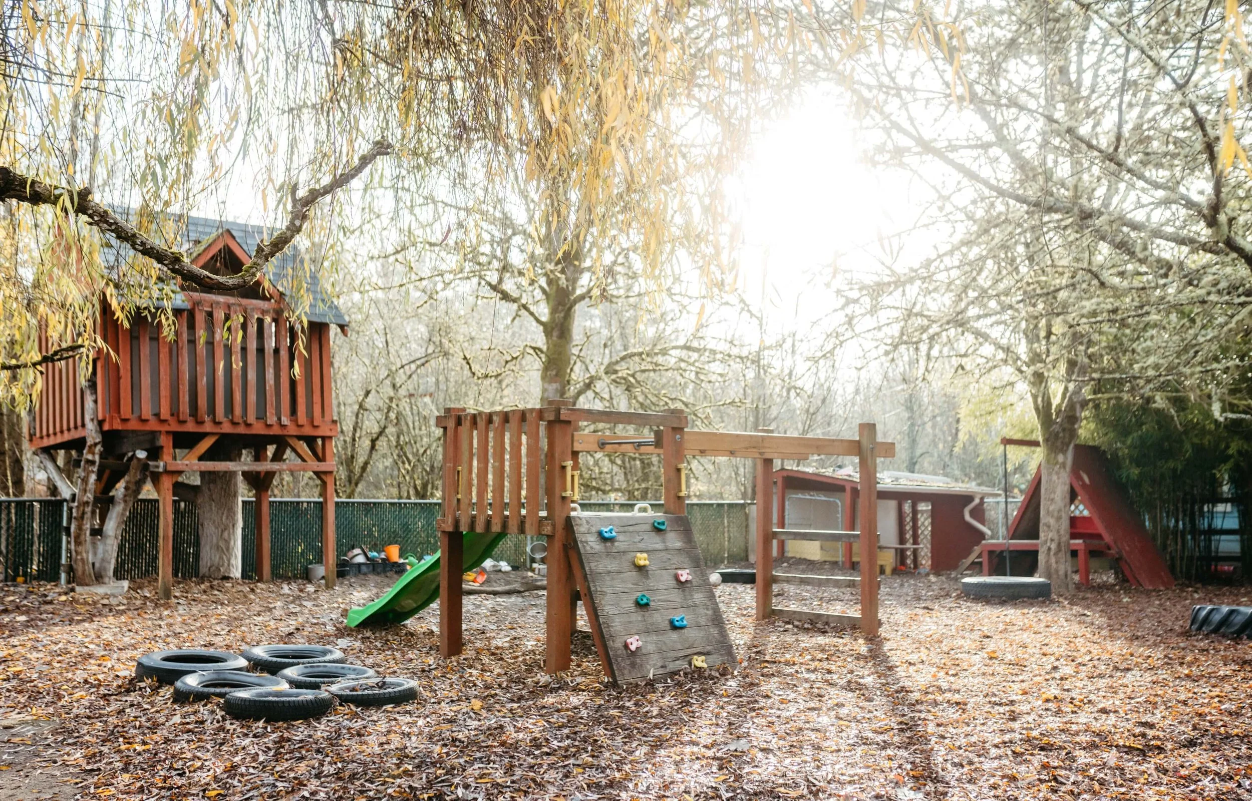 A playground with a slide, climbing wall, tire barriers, and a treehouse, surrounded by trees with fallen leaves on the ground, under sunlight.