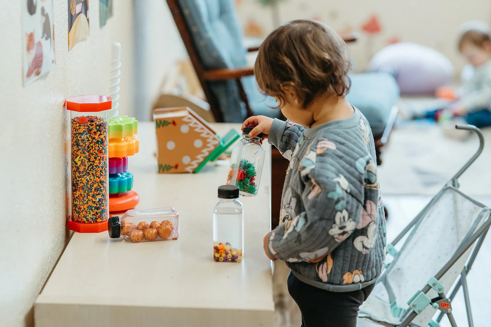 A young child with curly hair wearing a Mickey Mouse patterned jacket, looking at a small container with holiday decorations inside at a table.