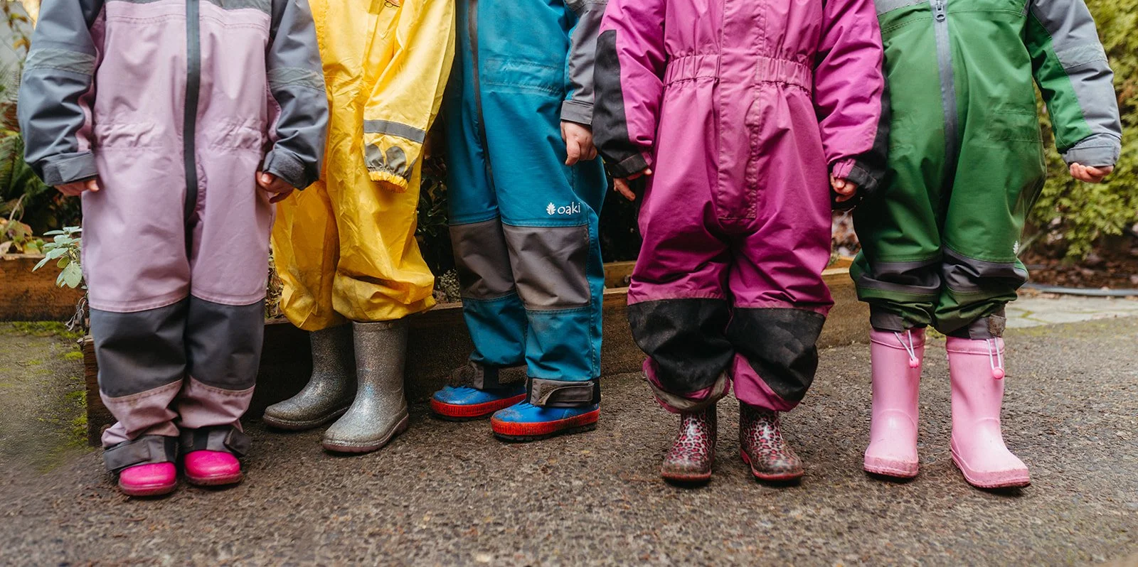 Children wearing colorful raincoats and rain boots standing outdoors on a wet ground.